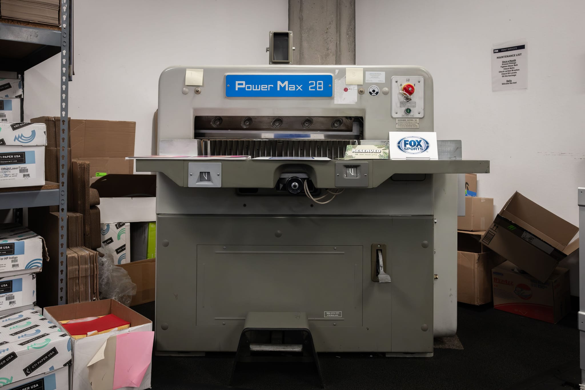 A large "Power Max 28" paper cutter with a FOX SPORTS logo in a cluttered workroom.