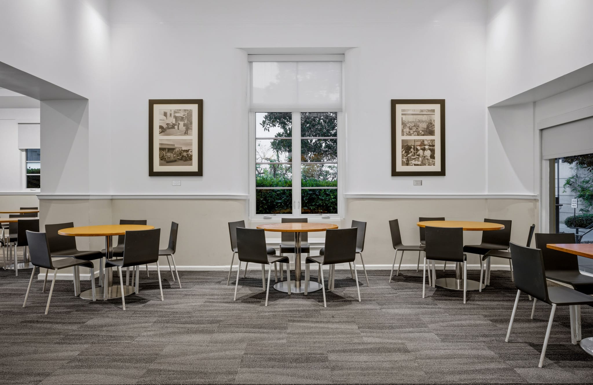 A bright, modern cafe or lounge with several round wooden tables, black chairs, a grey patterned carpet, white walls, a large window, and framed artwork.