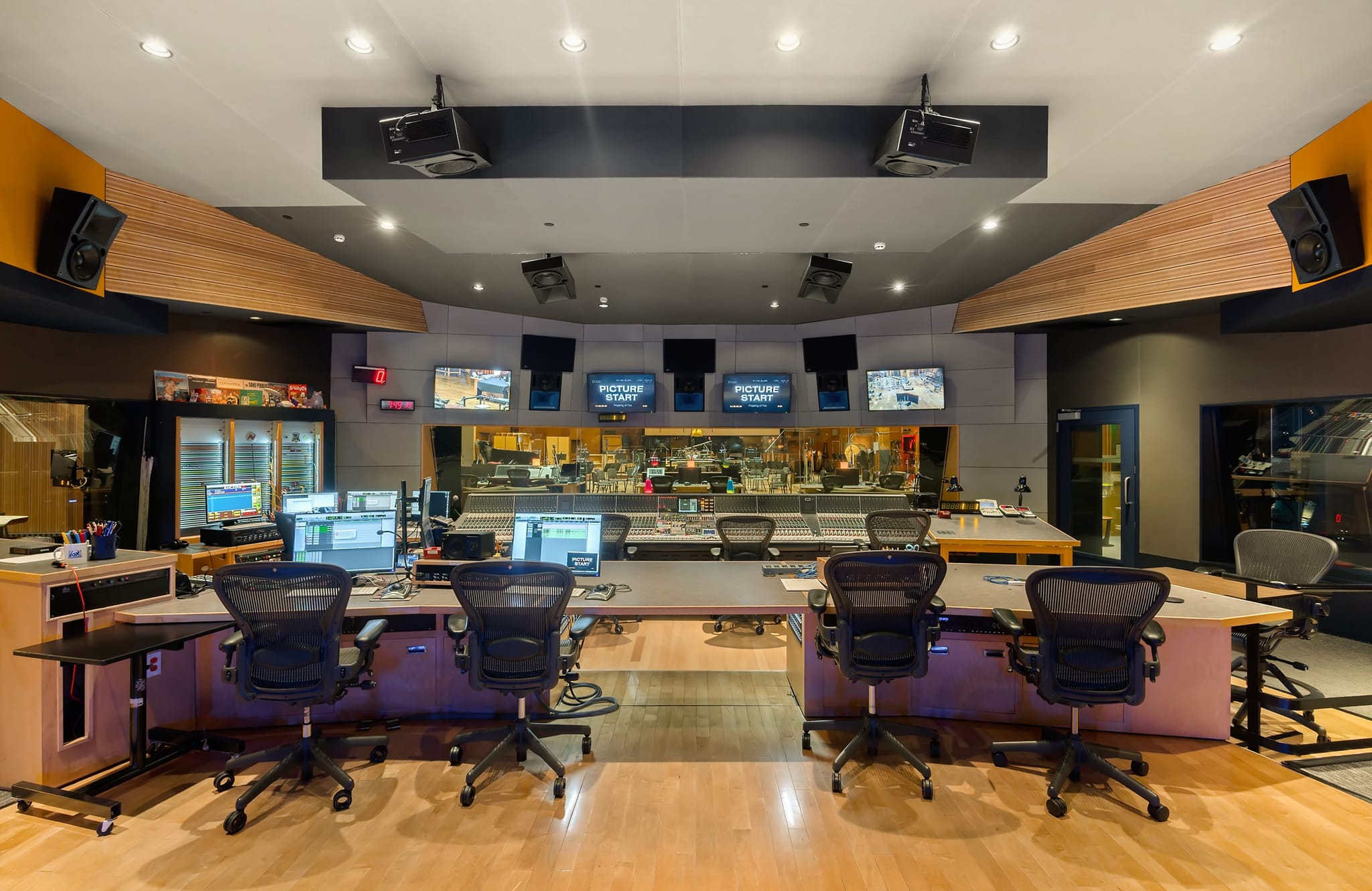 A modern studio control room with multiple workstations, computer monitors, and a large mixing console visible through a glass partition.