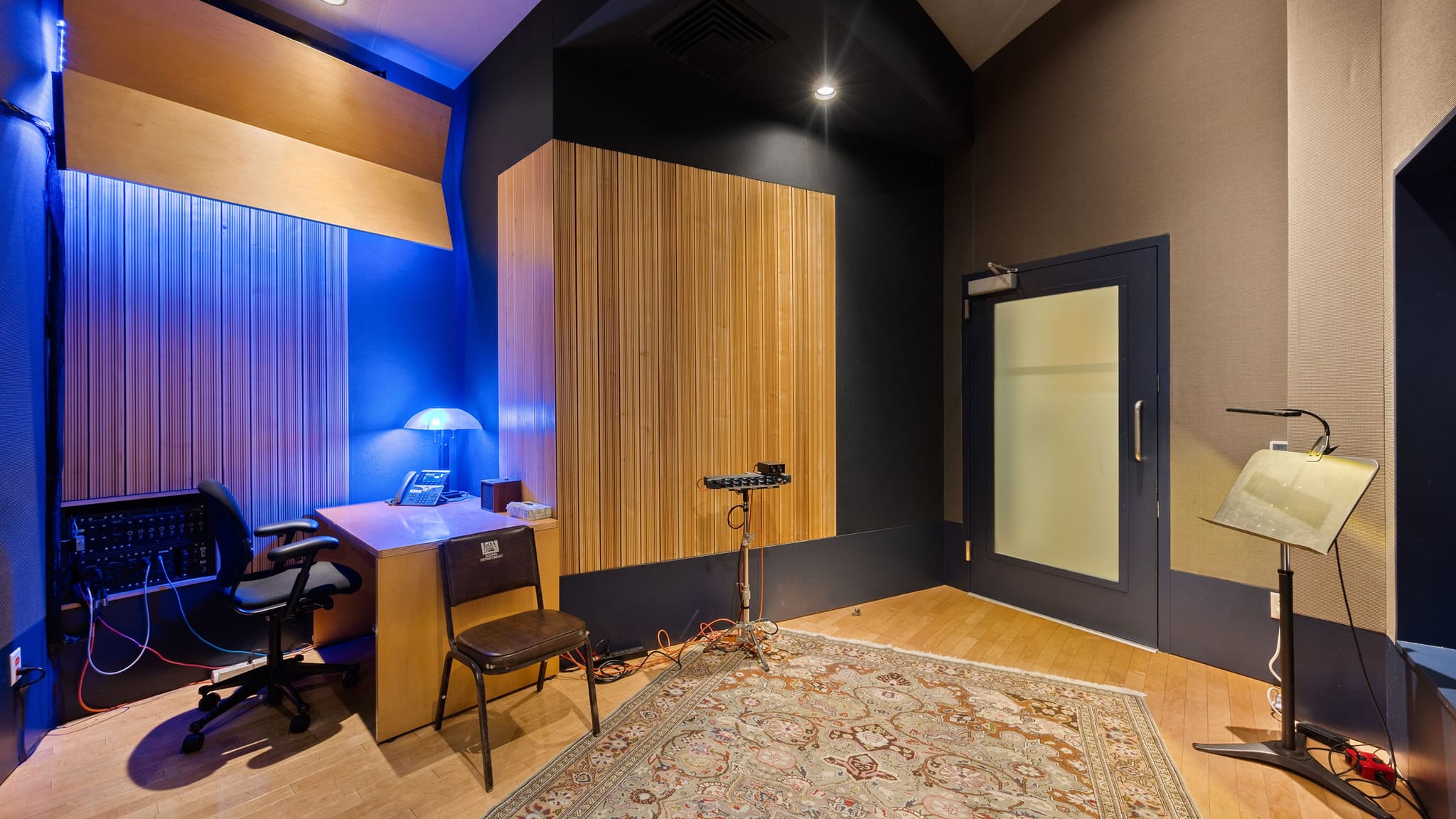 An empty recording studio with a desk, chairs, microphone, music stand, patterned rug, and sound-absorbing wall panels, some illuminated by blue light.