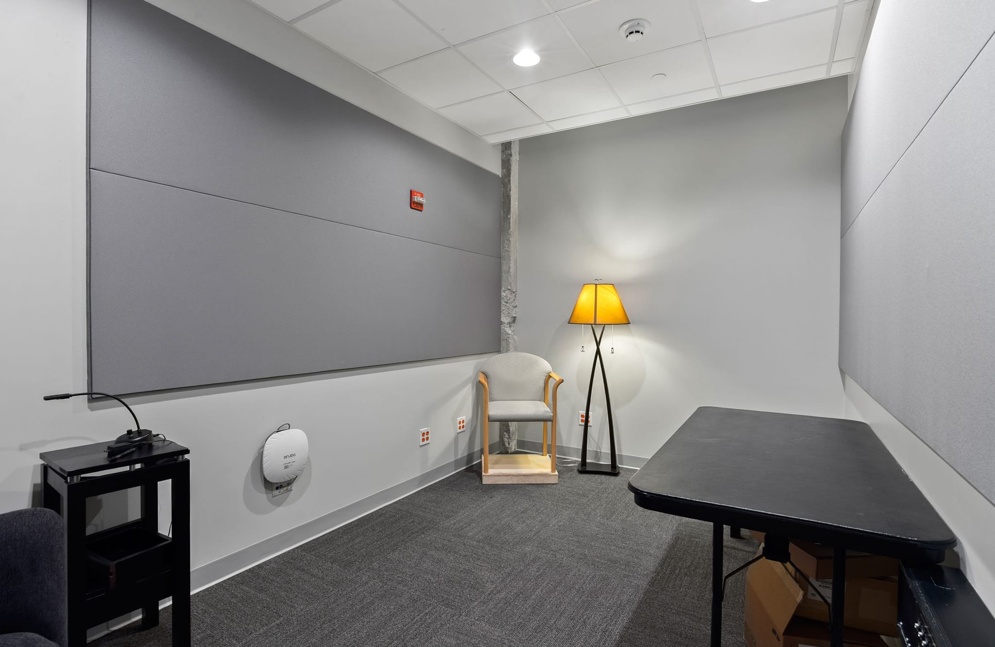 A room with grey acoustic panels on the walls, a microphone on a small black table, an armchair, a floor lamp with a yellow shade, and a black folding table.