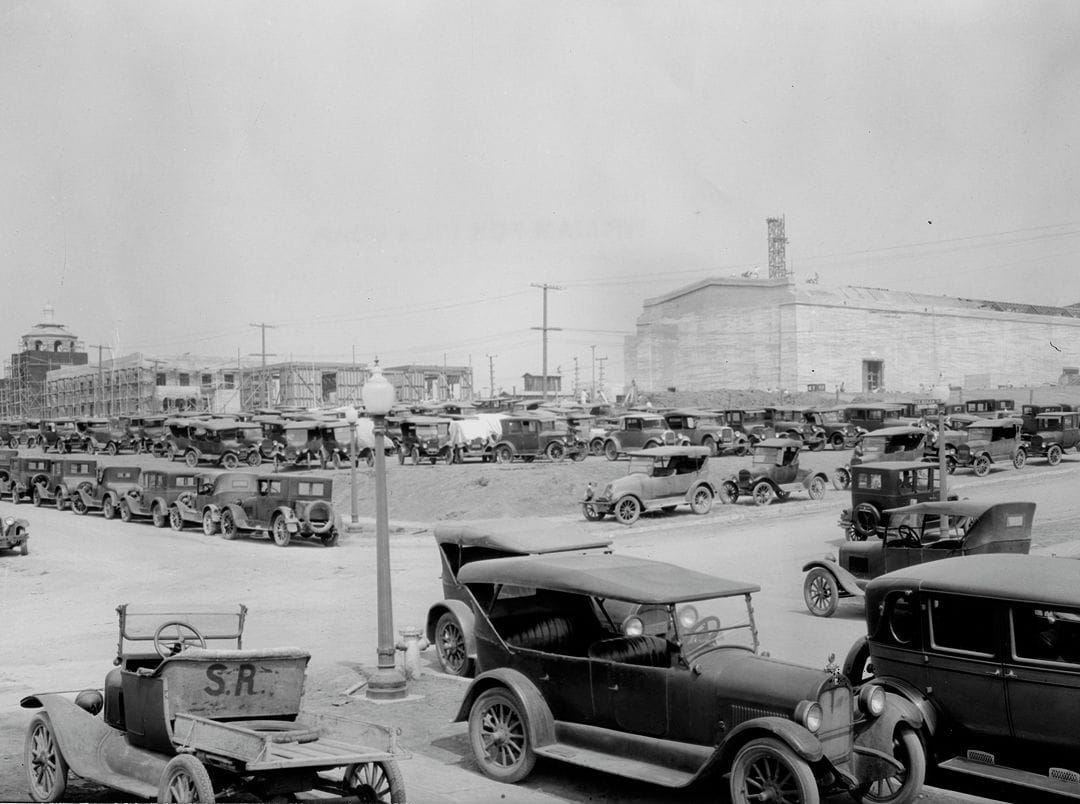 Black and white image of many 1920s-era cars parked in an open area, with a large building and construction sites in the background.