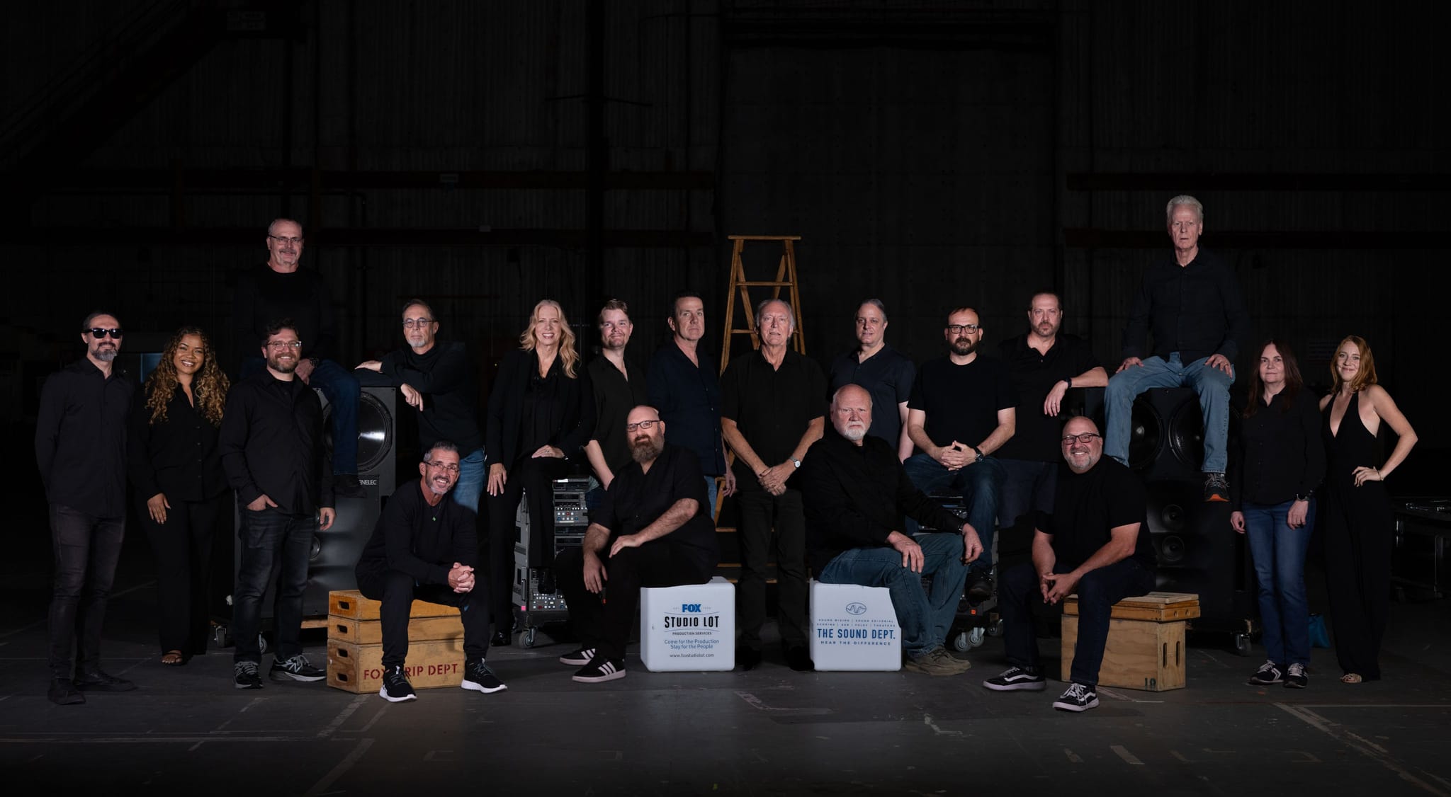 A large group of people in dark clothing poses in a studio with production equipment and branded boxes.