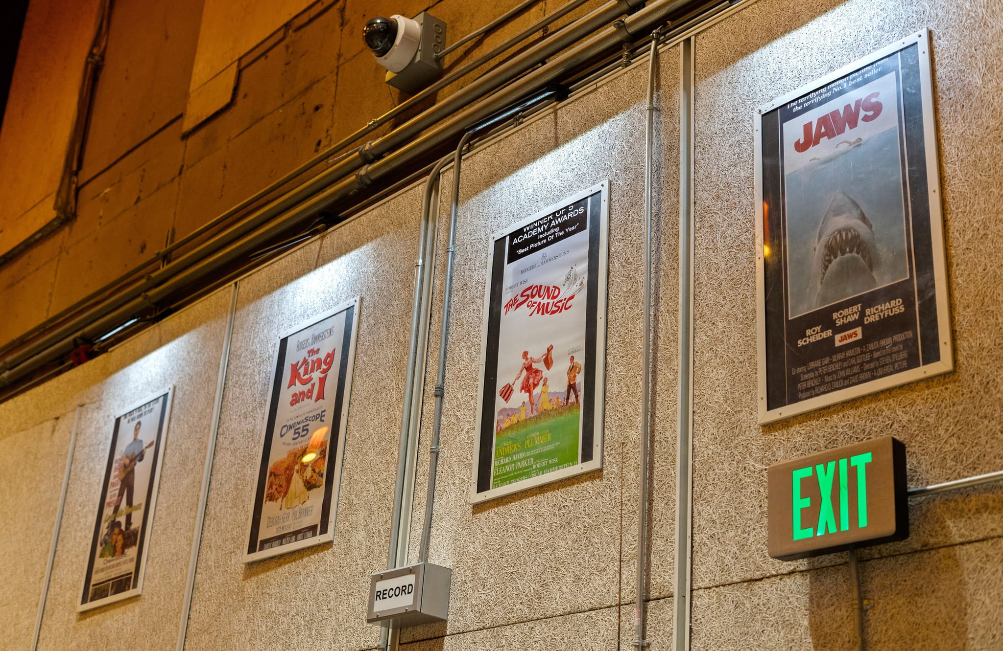 Movie posters for "Jaws," "The Sound of Music," and "The King and I" on a wall with an illuminated "EXIT" sign and a security camera.