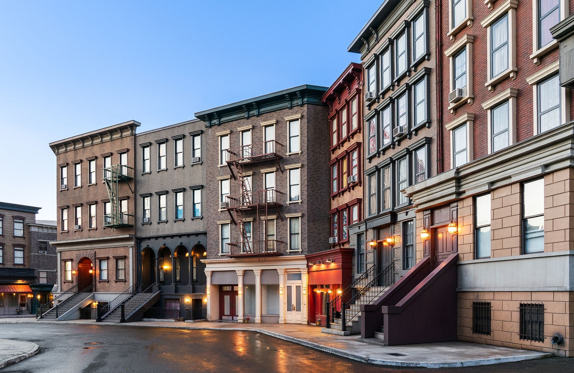 Old brick buildings line a wet city street at dusk, with lights illuminating the facades.