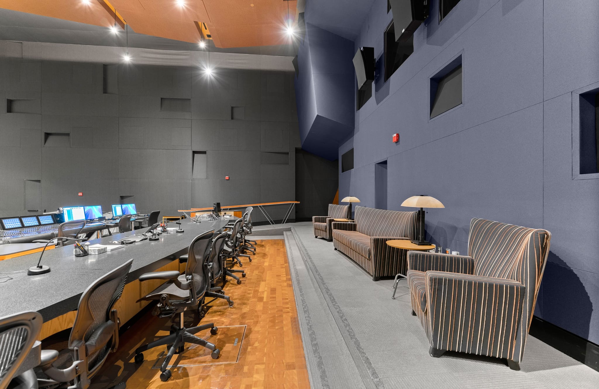 Modern control room with a long table of monitors and chairs, adjacent to a lounge area with striped sofas and armchairs.