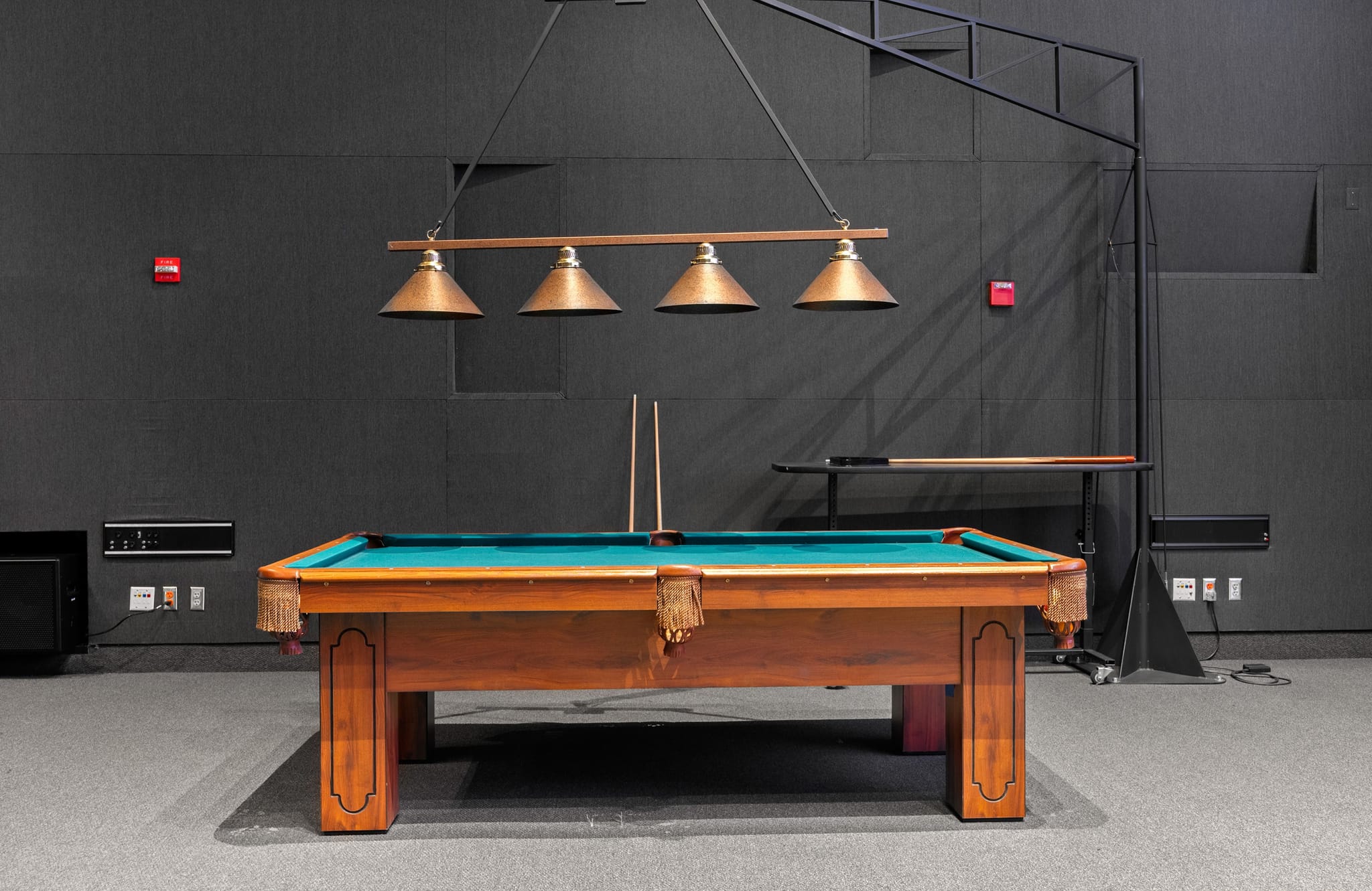 A wooden billiards table with teal felt, two cues, and an overhead four-lamp bronze light fixture in a dark gray room.