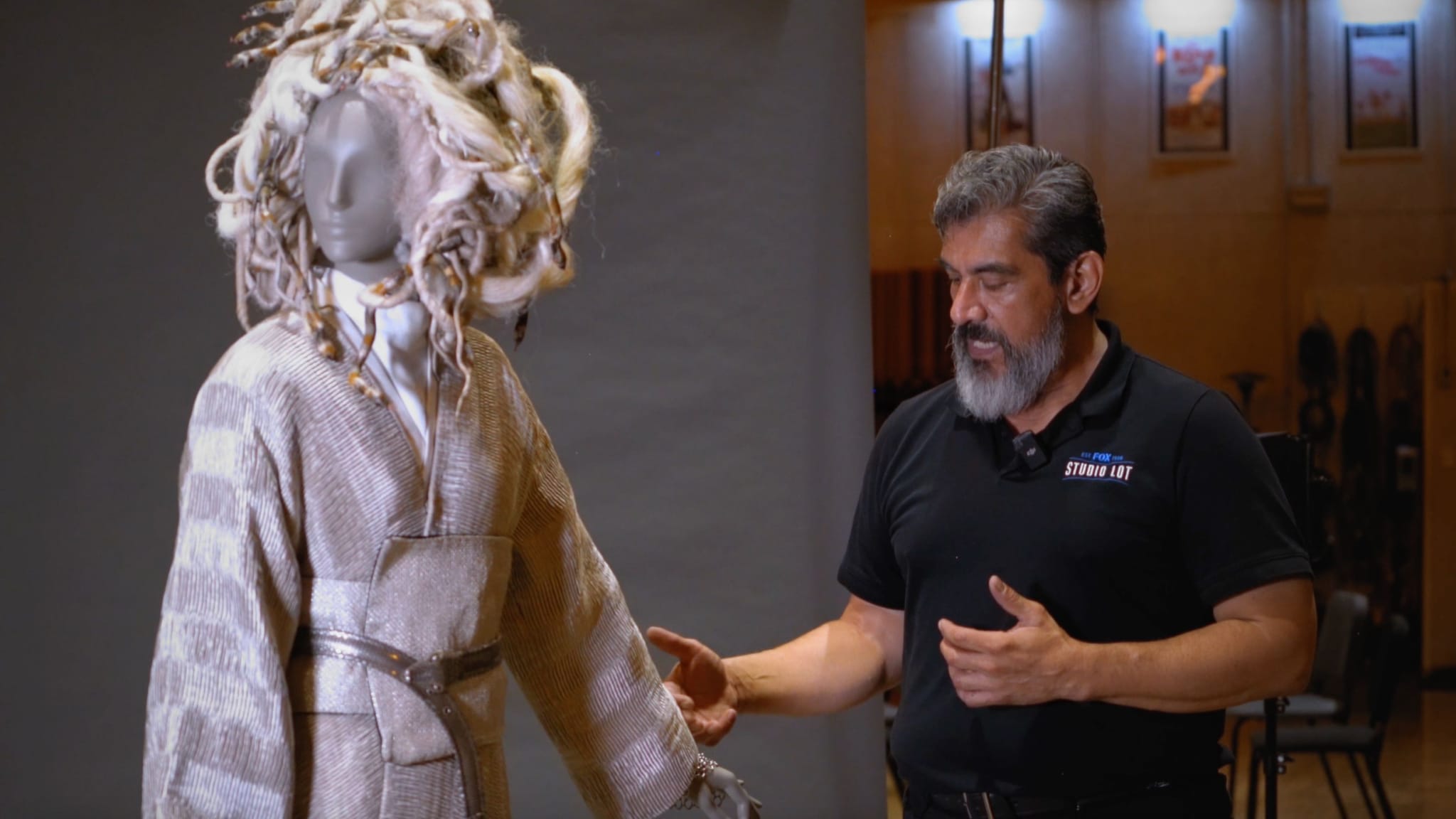 A man in a black shirt gestures toward a mannequin dressed in an elaborate light-colored fantasy costume with a distinctive wig.