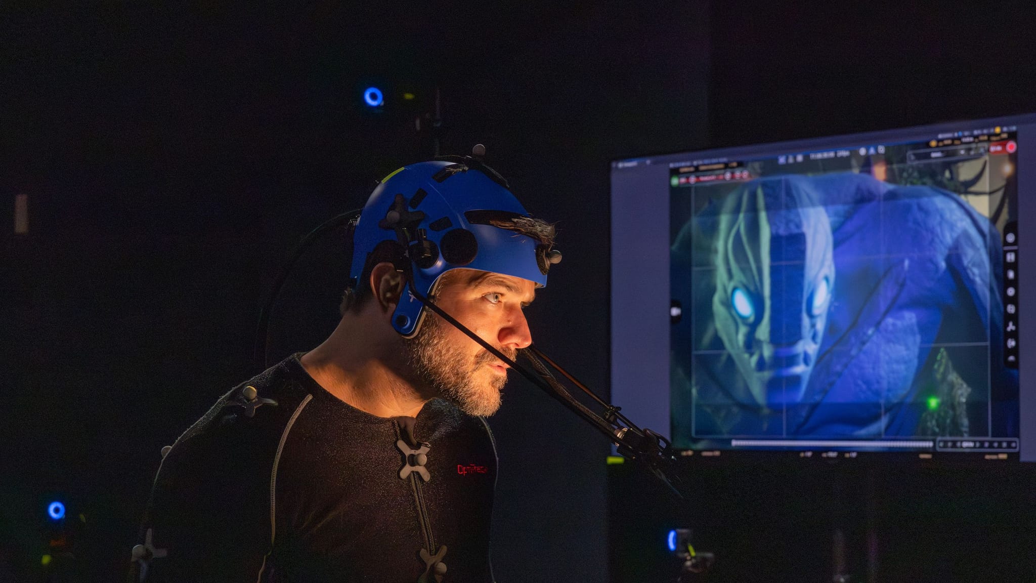 Man in motion capture gear looks at a monitor displaying a blue alien character with glowing eyes.
