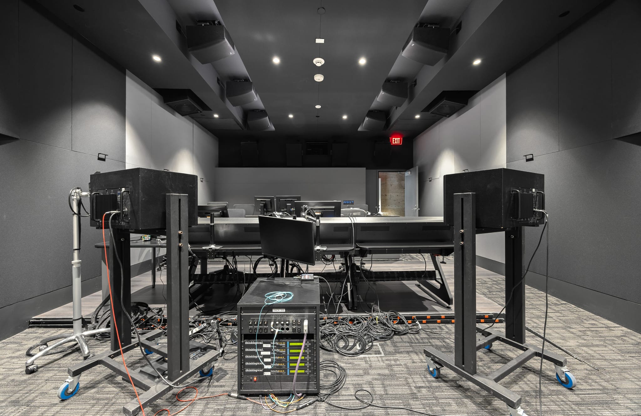 A dark, modern audio control room or studio with large speakers on stands, a central mixer unit, and multiple computer workstations in the background.