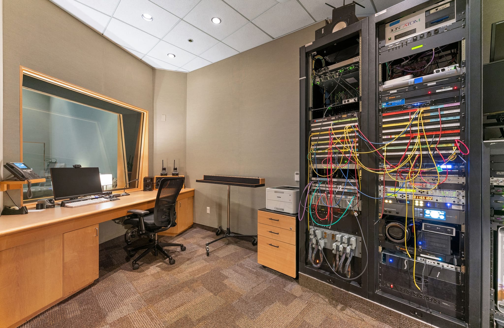 A modern control room with a desk, computer, a large viewing window, and two tall racks filled with electronic gear.