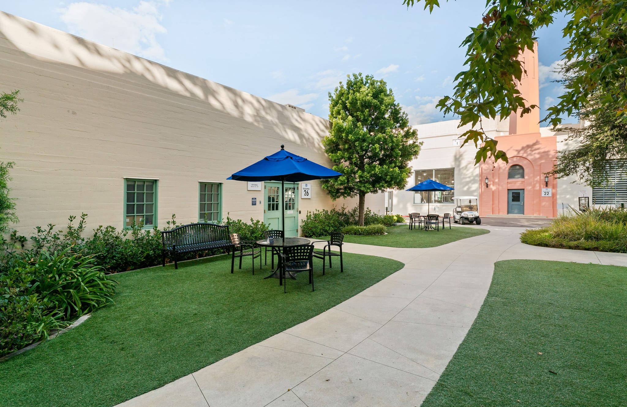 Courtyard with artificial turf, winding paths, and outdoor seating under blue umbrellas, framed by buildings and trees.