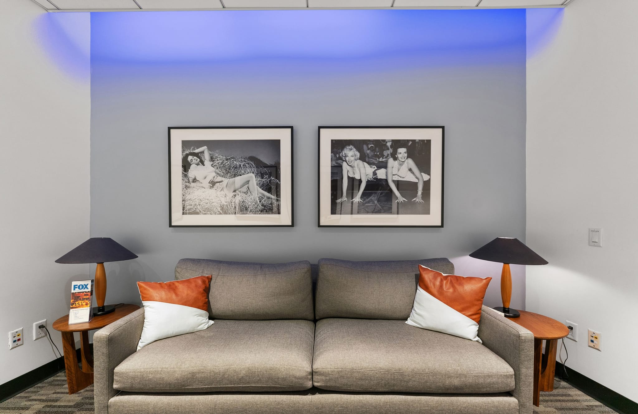 A gray couch with decorative pillows, flanked by end tables with lamps, sits beneath two framed black and white photos of women, with blue accent lighting above.