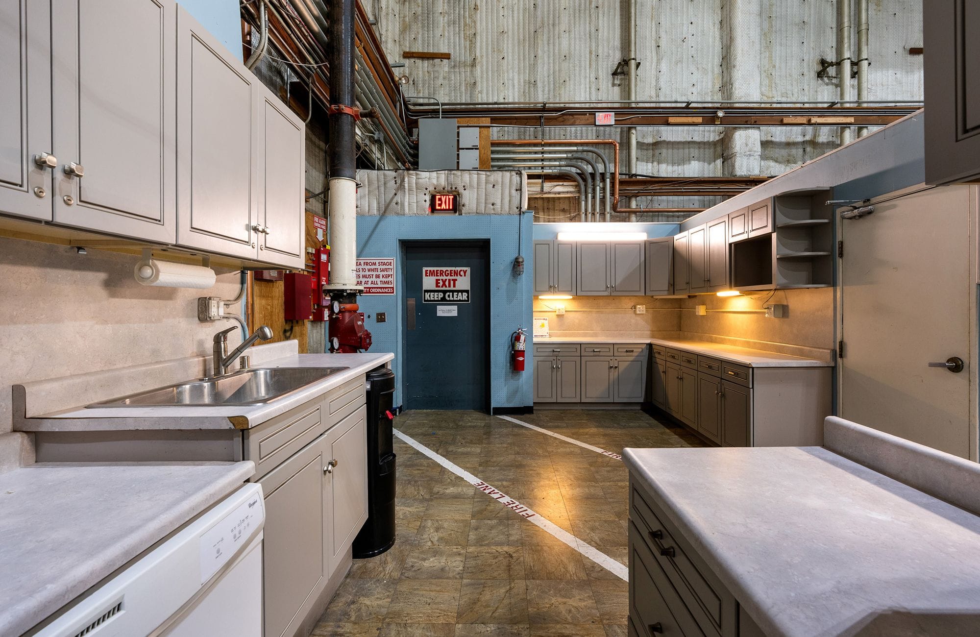 A kitchen area with gray cabinets and counters, including a sink and under-cabinet lighting, set within a large industrial or warehouse space with an emergency exit.