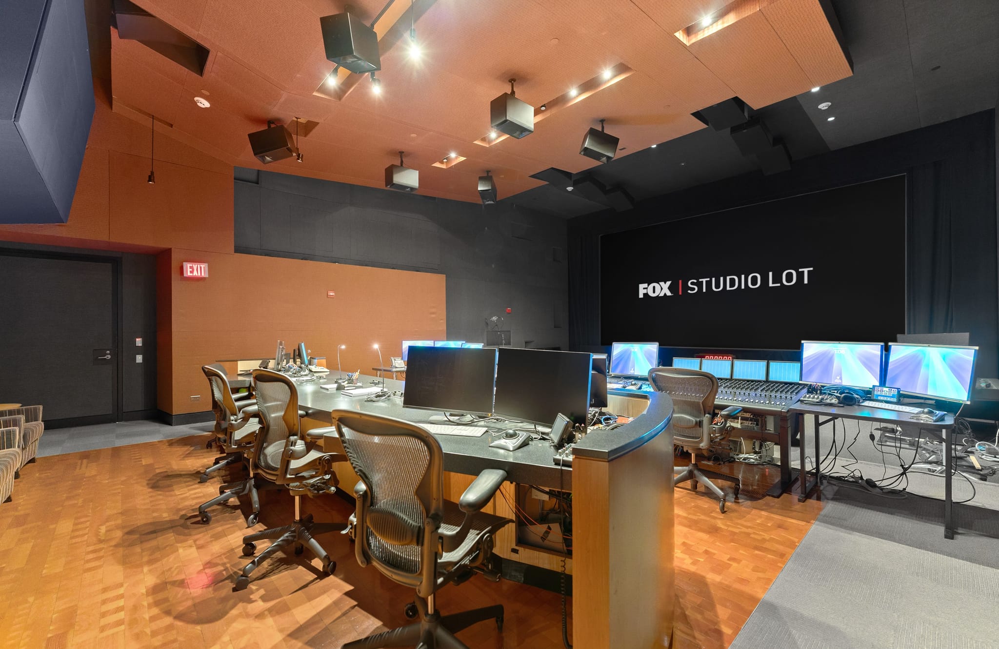 A modern media production studio control room with multiple workstations, monitors, a sound mixing console, and a large screen displaying "FOX | STUDIO LOT."
