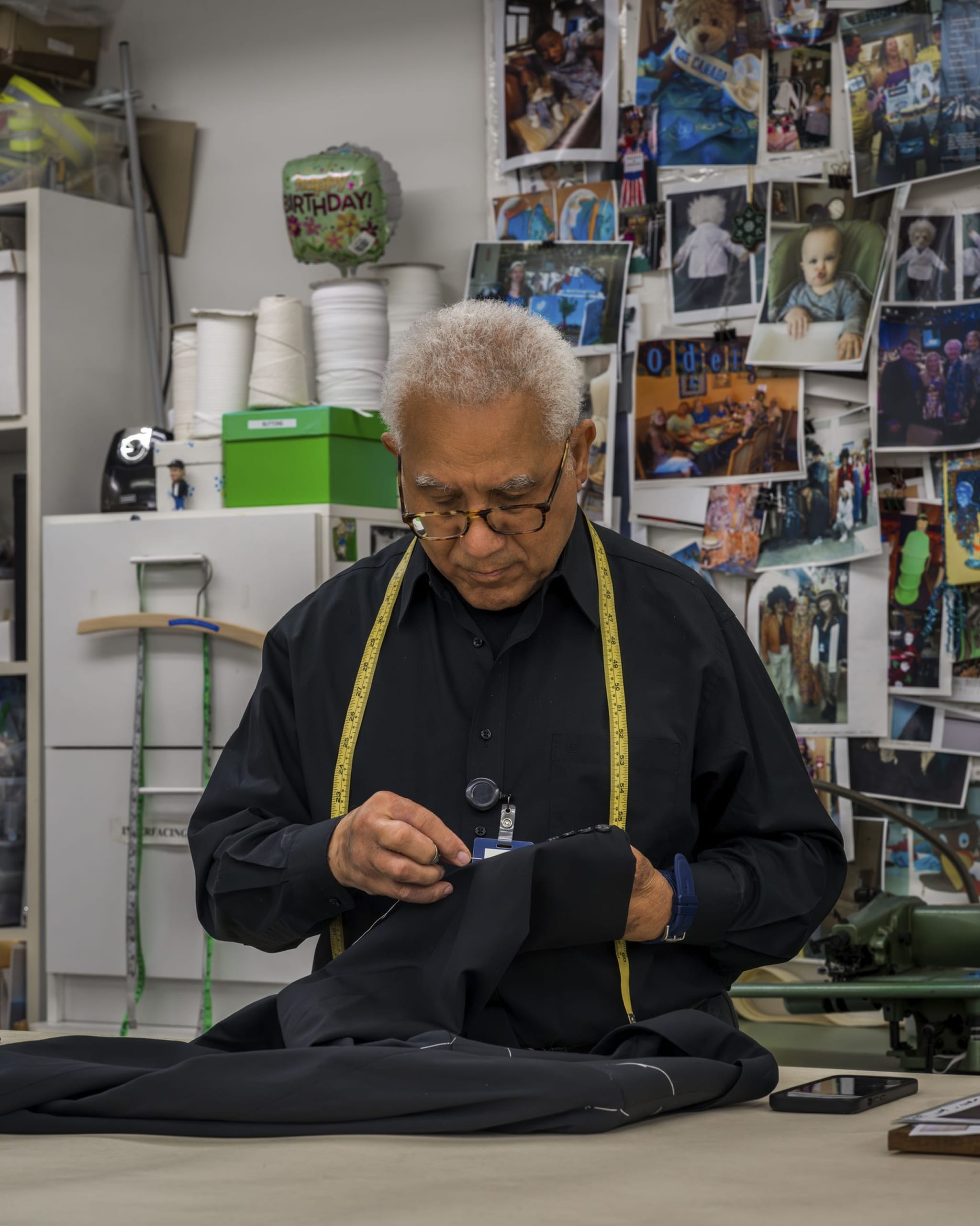 An older man with a measuring tape around his neck hand-sews black fabric in a workshop.