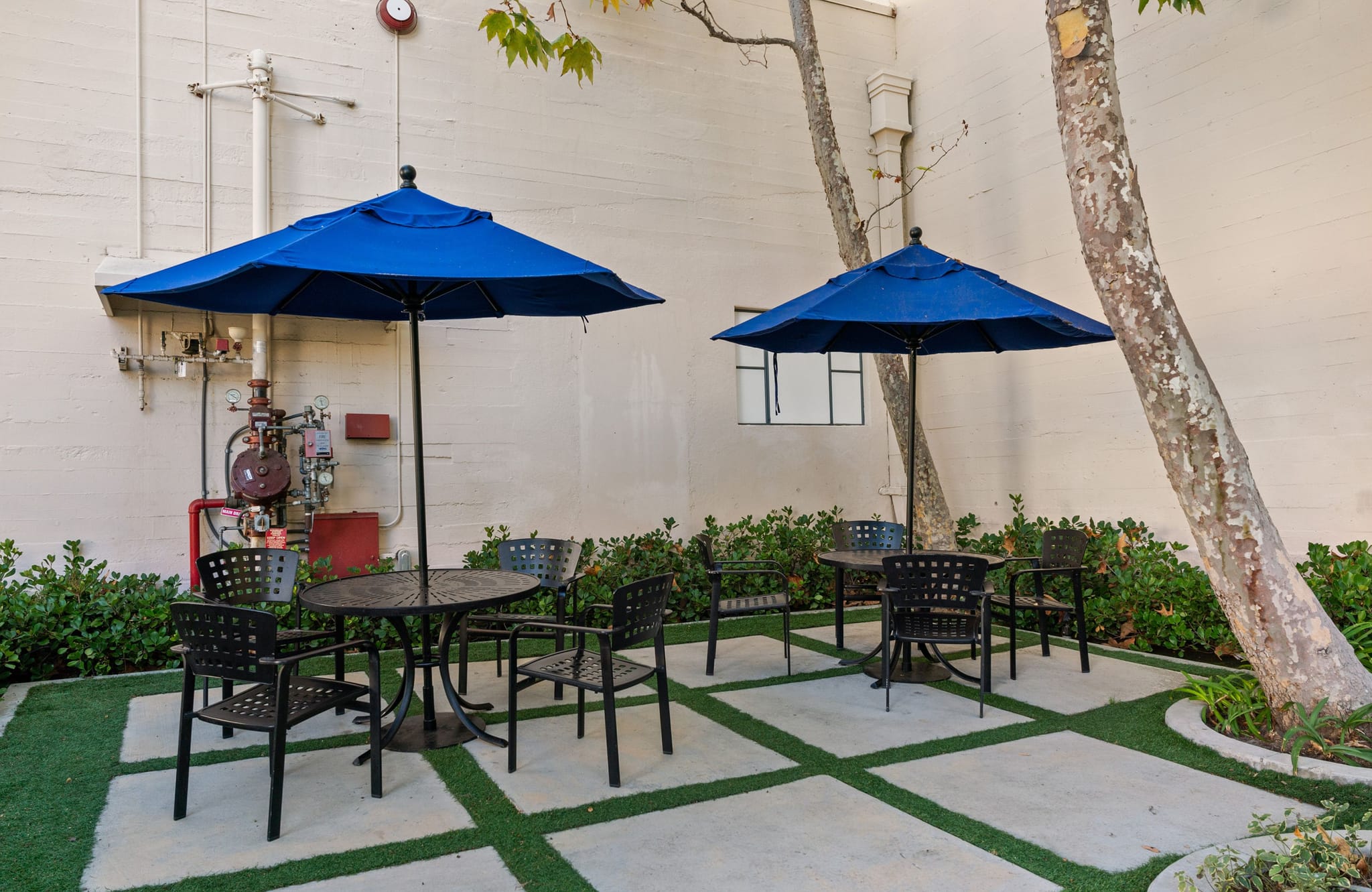An outdoor patio with two blue umbrellas over tables and chairs, set on a grid of concrete pavers and green turf, surrounded by foliage.