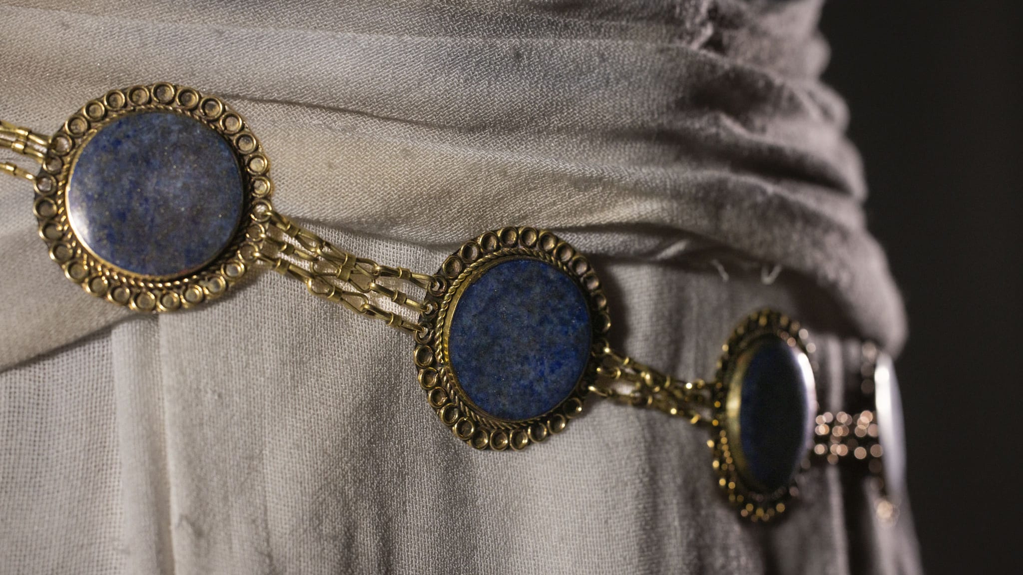 Decorative gold belt with three circular lapis lazuli stones over light fabric.