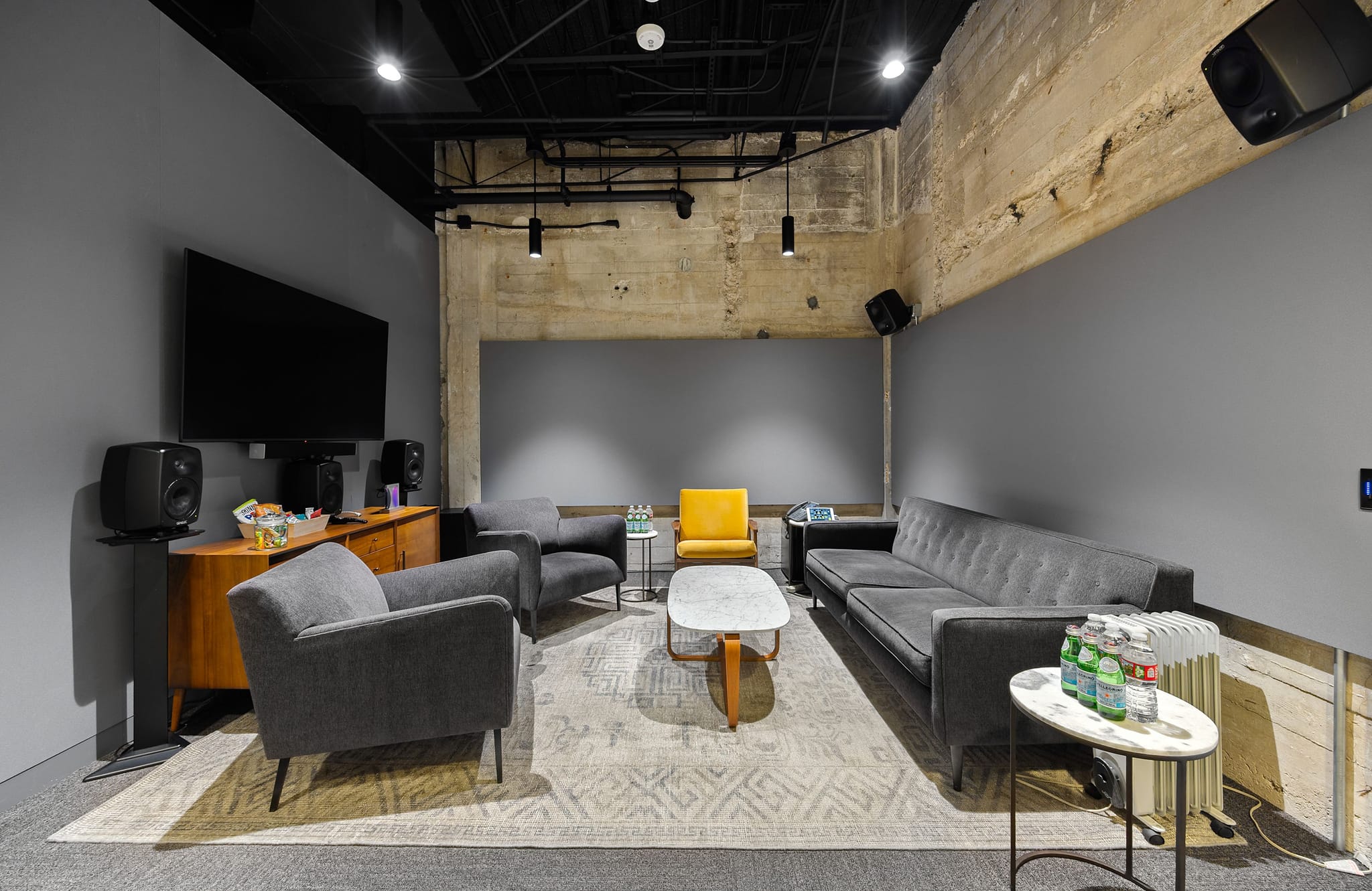 A modern media lounge with gray walls, exposed concrete, a large TV and speakers, gray sofas, armchairs, and yellow accent chairs around a marble coffee table.