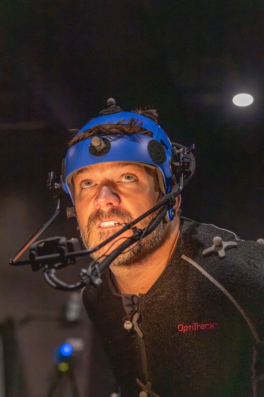 A man in a blue motion capture helmet and dark OptiTrack suit looks up.
