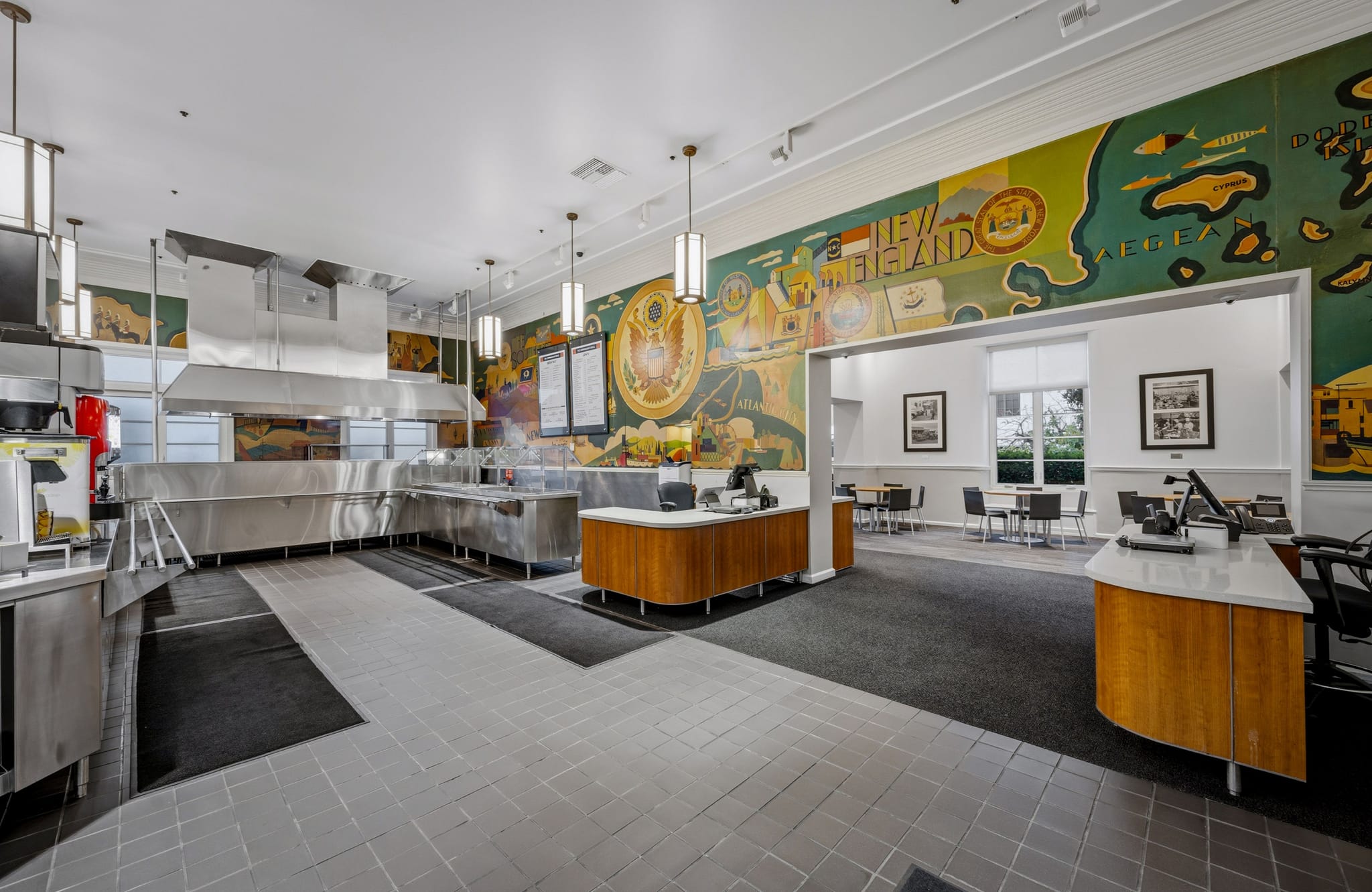 Spacious cafeteria with stainless steel serving line, dining tables, and colorful New England-themed murals on the walls.