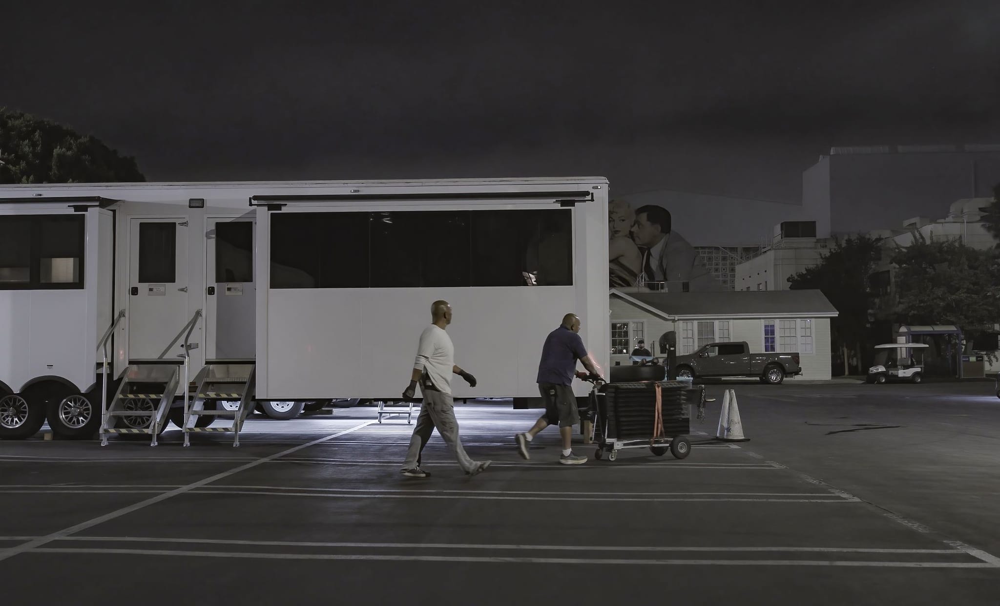Nighttime film set with a large white trailer, crew members, and a giant kissing couple image in the background.