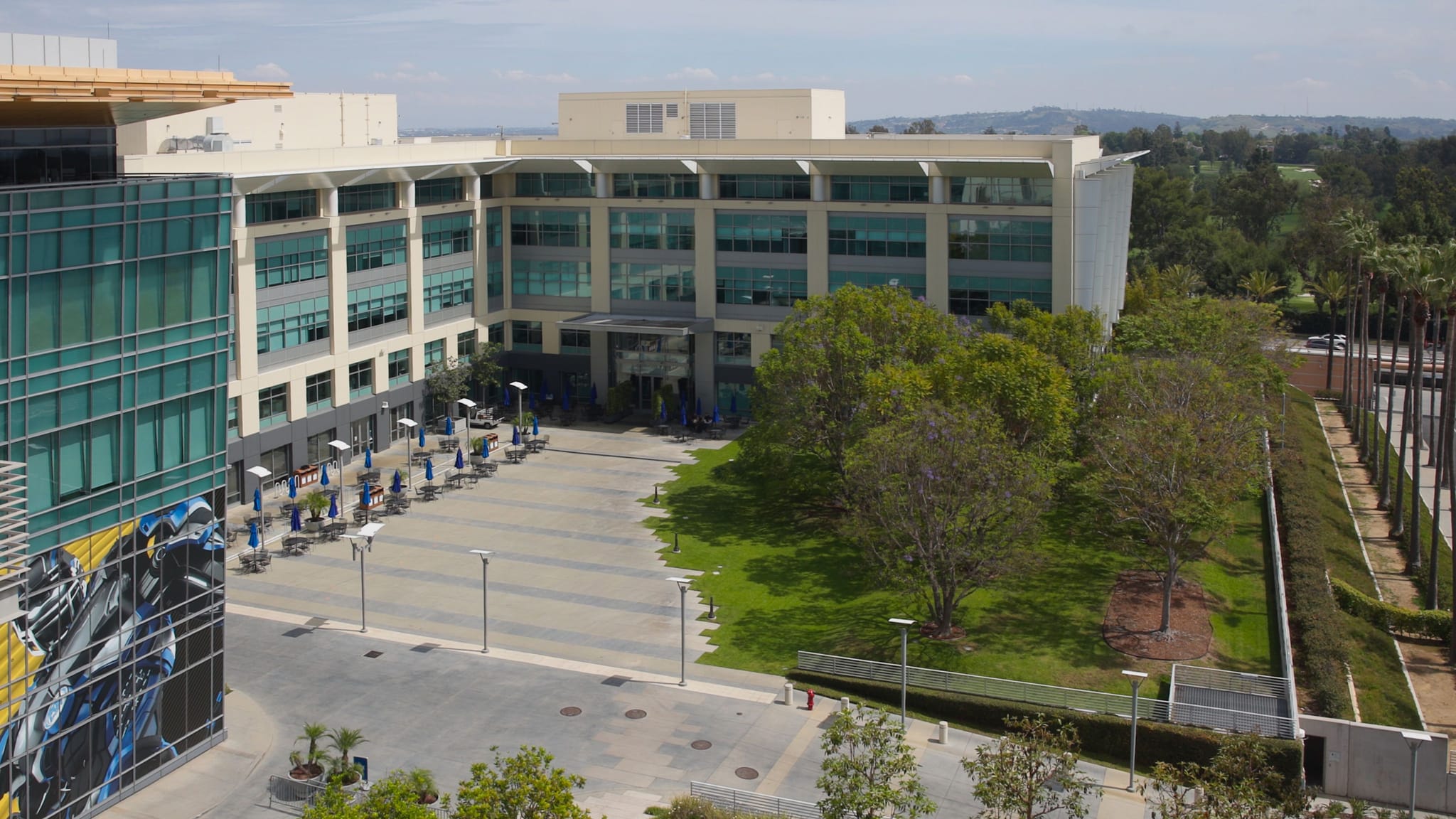Modern office complex with an outdoor plaza, patio seating, trees, and a colorful mural on a glass building.