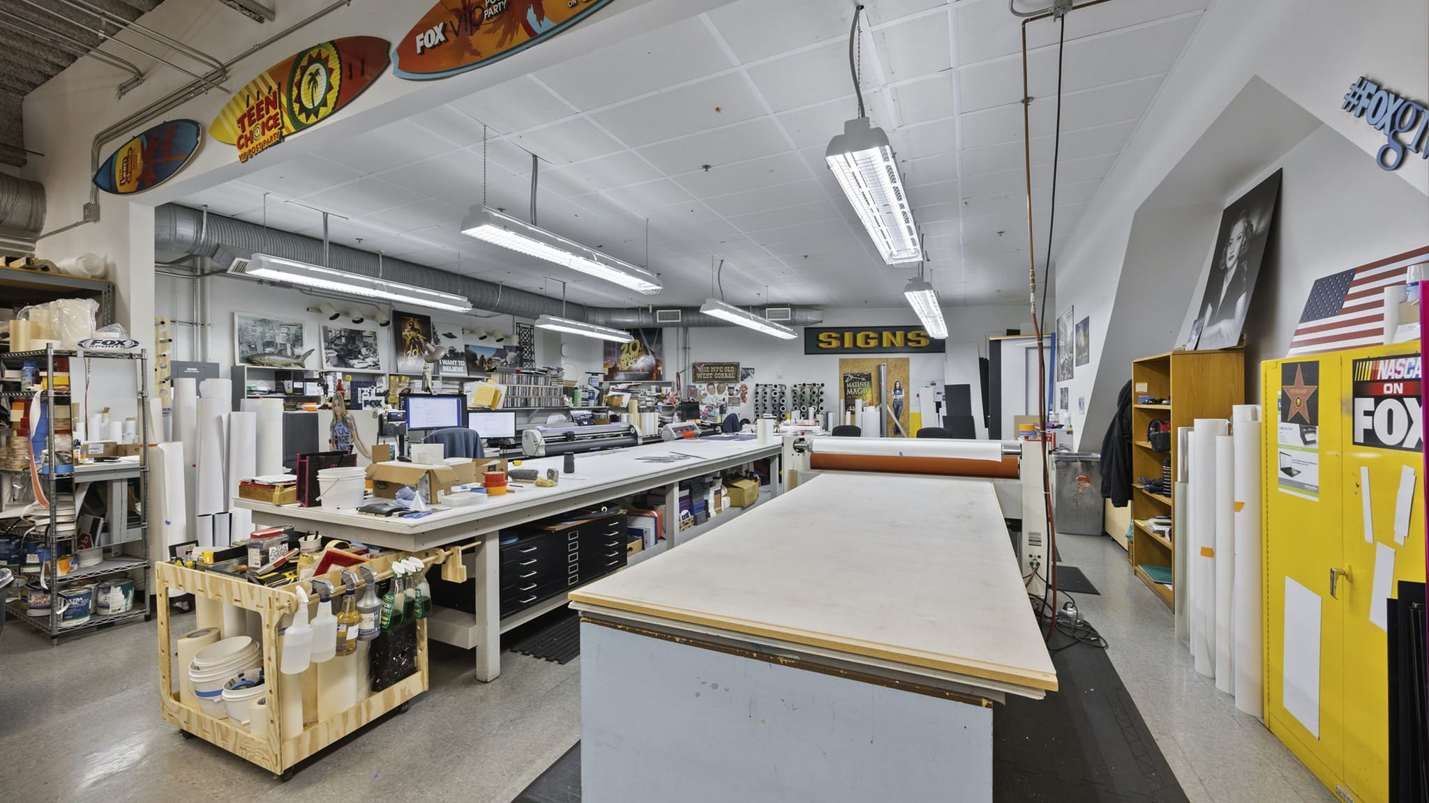 An expansive sign-making and print workshop with multiple work tables, rolls of material, computers, and branded surfboards hanging from the ceiling.