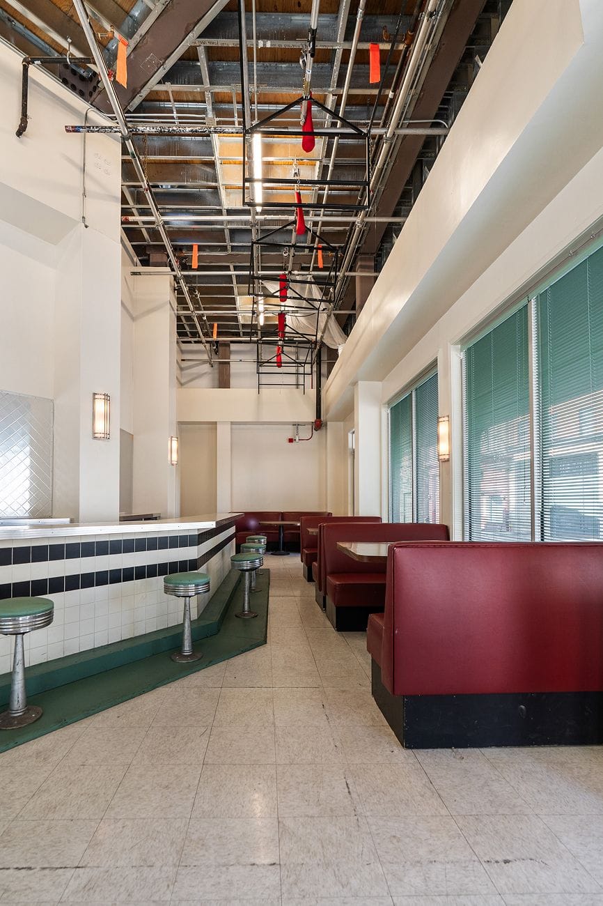 A retro-style diner interior with a counter and green stools on the left, red booths on the right, and an exposed industrial ceiling.
