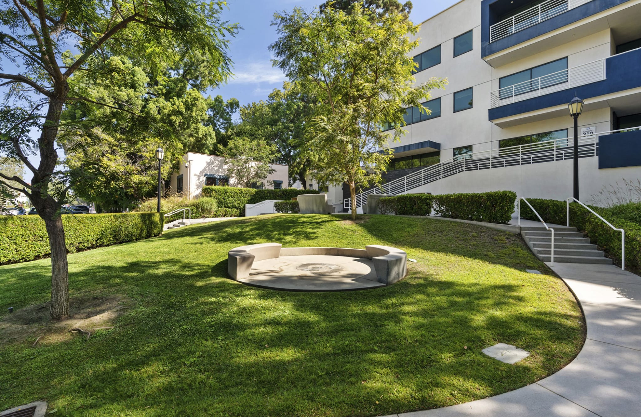 Green lawn with trees, hedges, a circular concrete seating area, and a modern white and blue building with stairs.