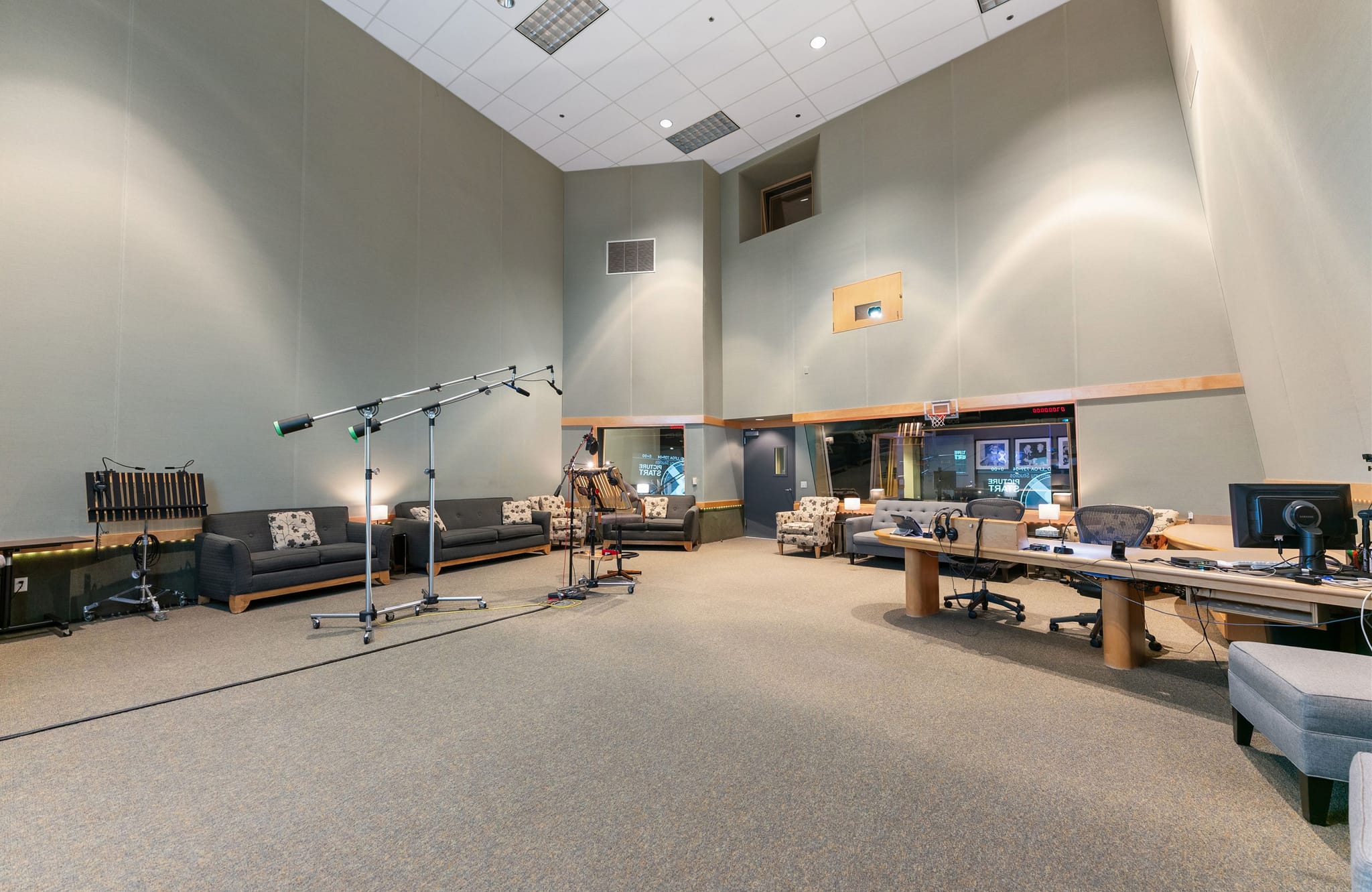 A spacious recording studio with multiple microphone stands, sofas, a control room visible through glass, and a large mixing desk with computer monitors.