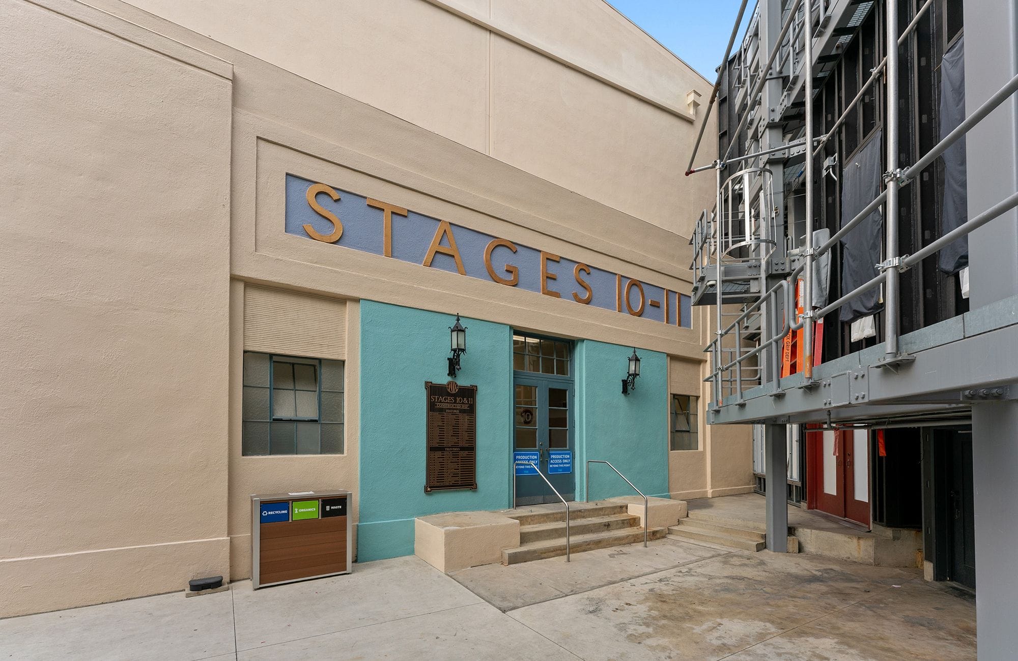 Exterior view of a building with "STAGES 10-11" on its facade, above a turquoise entrance with steps, next to a metal scaffolding structure.