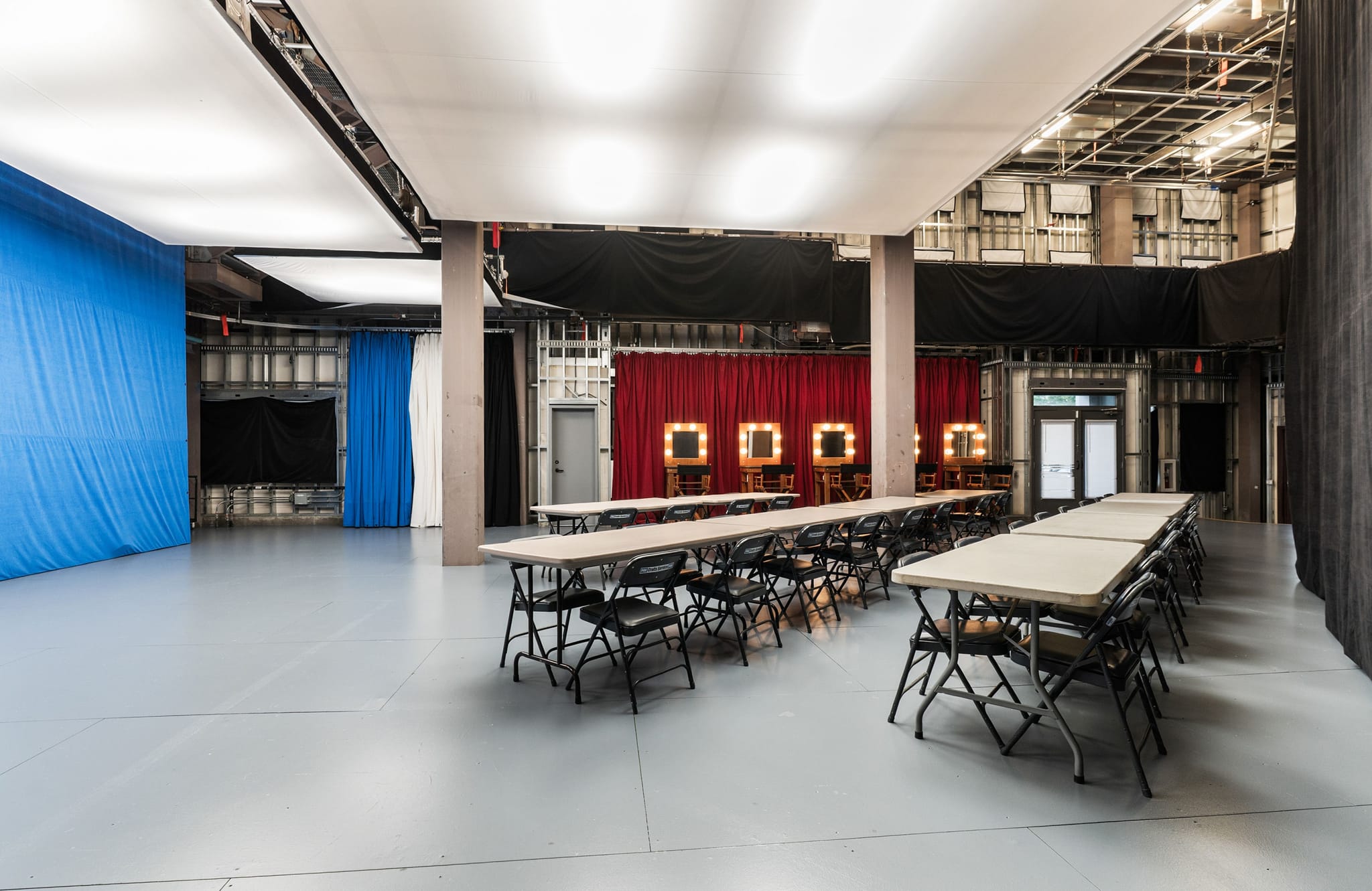 A spacious studio with a blue screen, rows of tables and chairs, and illuminated vanity mirrors against red curtains.