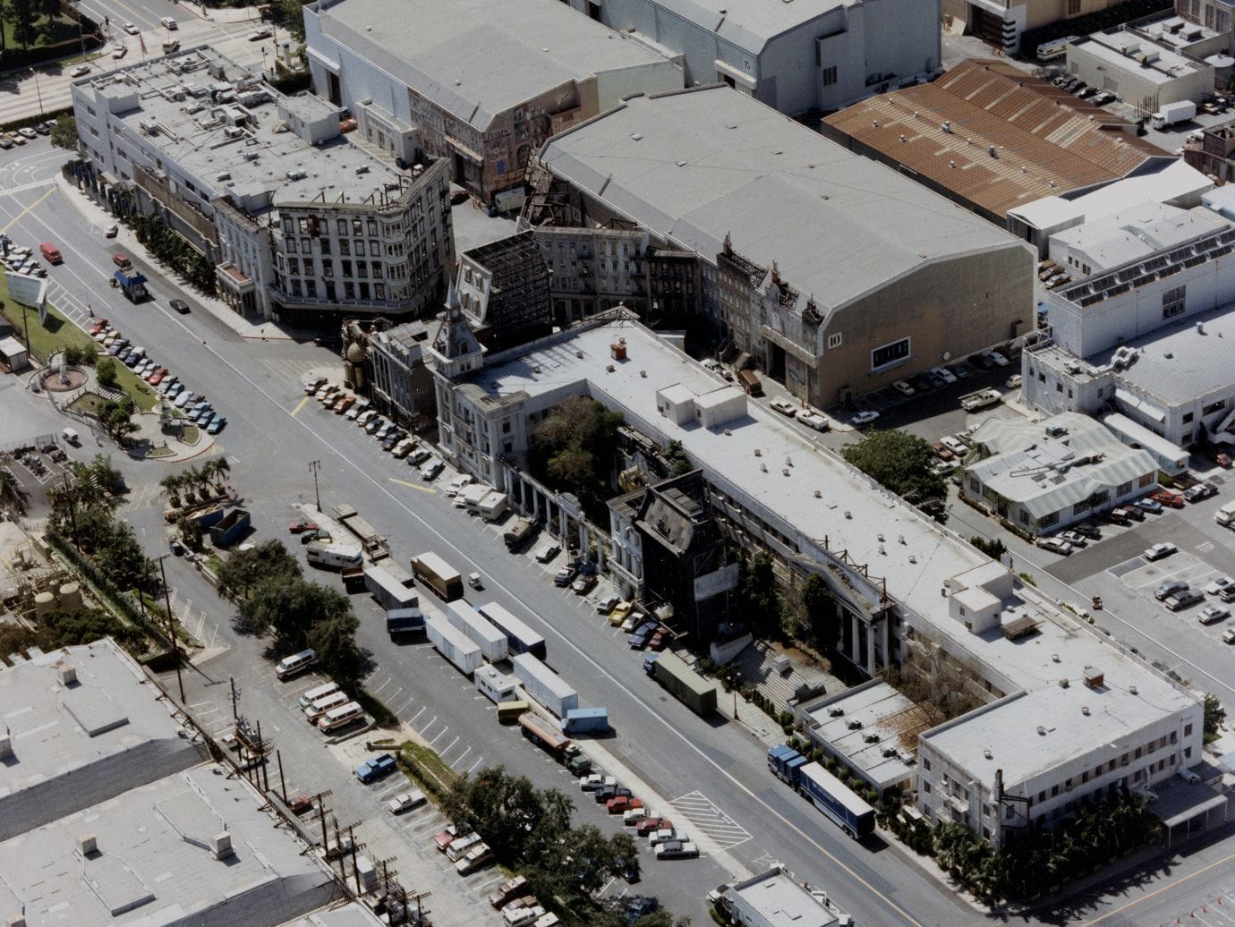 Aerial view of a film studio lot with ornate building facades, sound stages, streets, and parking lots filled with cars and trucks.
