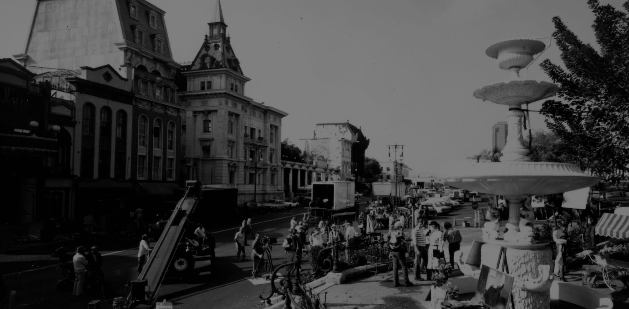 Black and white photograph of a street scene with classic buildings, a multi-tiered fountain, and many people.