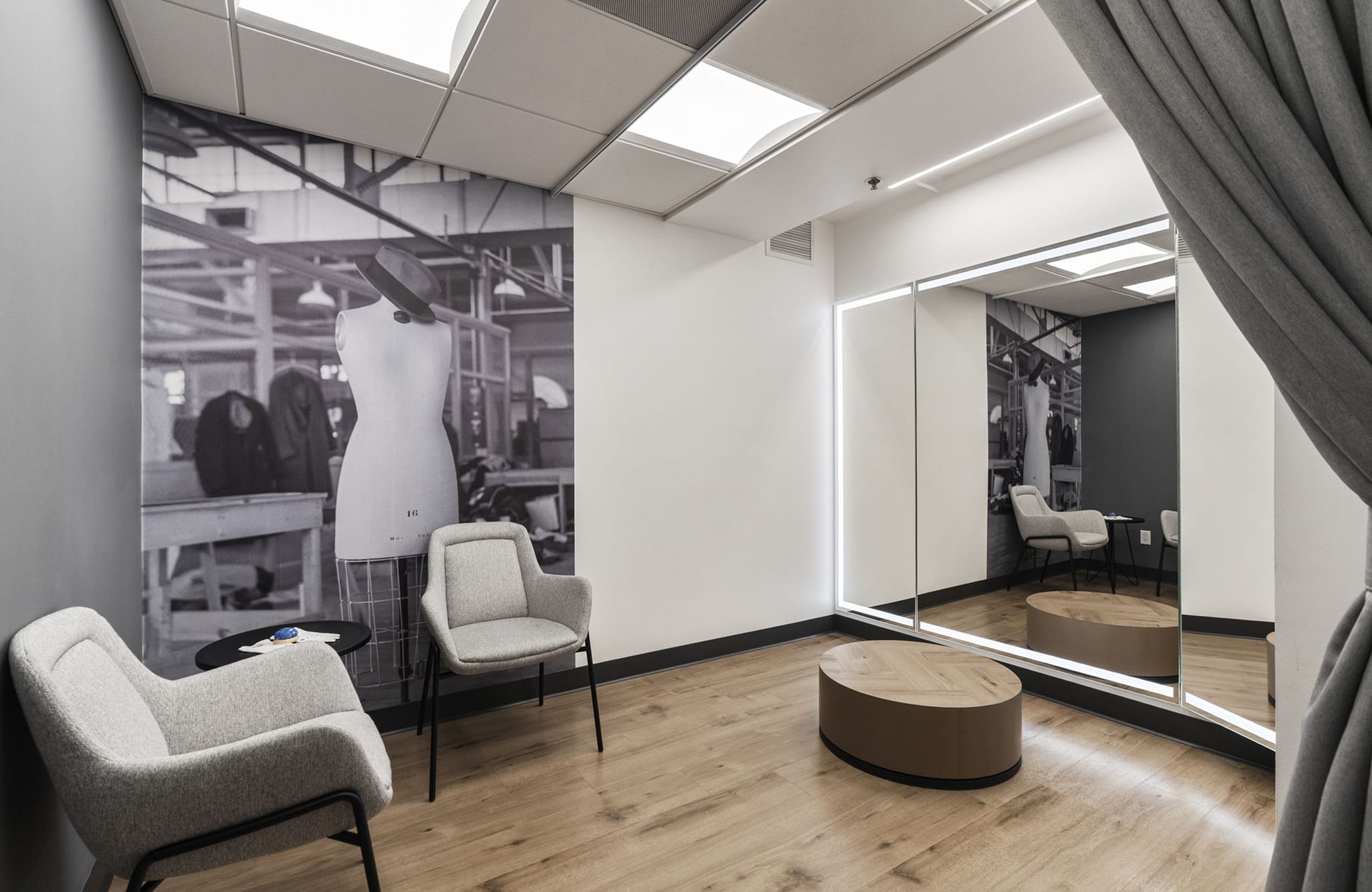 Modern fitting room with a large mirror framed by LED lights, an oval wooden bench, two grey armchairs, and a black and white mural of a garment factory.