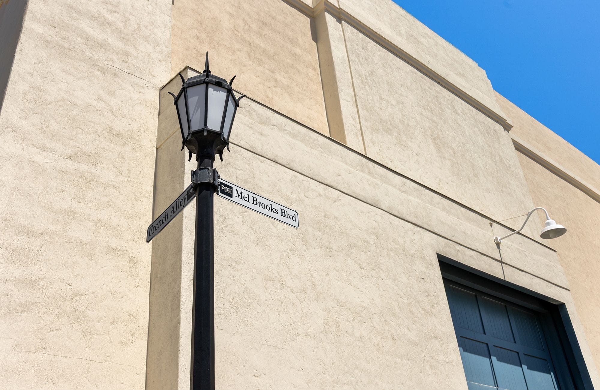 Black street lamp post with "French Alley" and "Mel Brooks Blvd" signs against a beige stucco building under a blue sky.