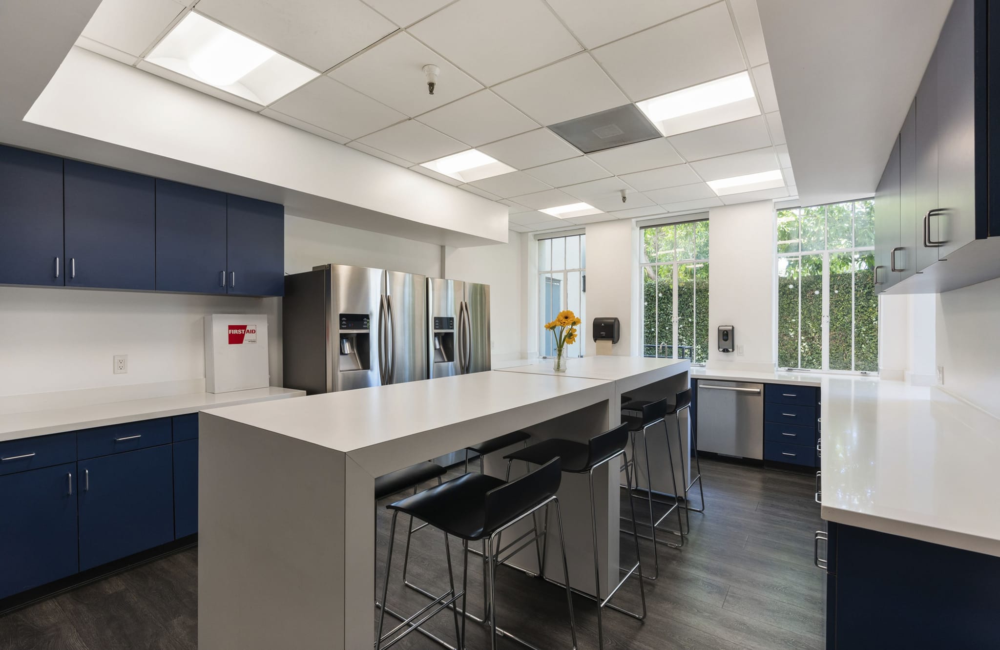 A bright, modern office kitchen featuring navy blue cabinets, white countertops, three stainless steel refrigerators, a large island with black bar stools, and windows overlooking green foliage.