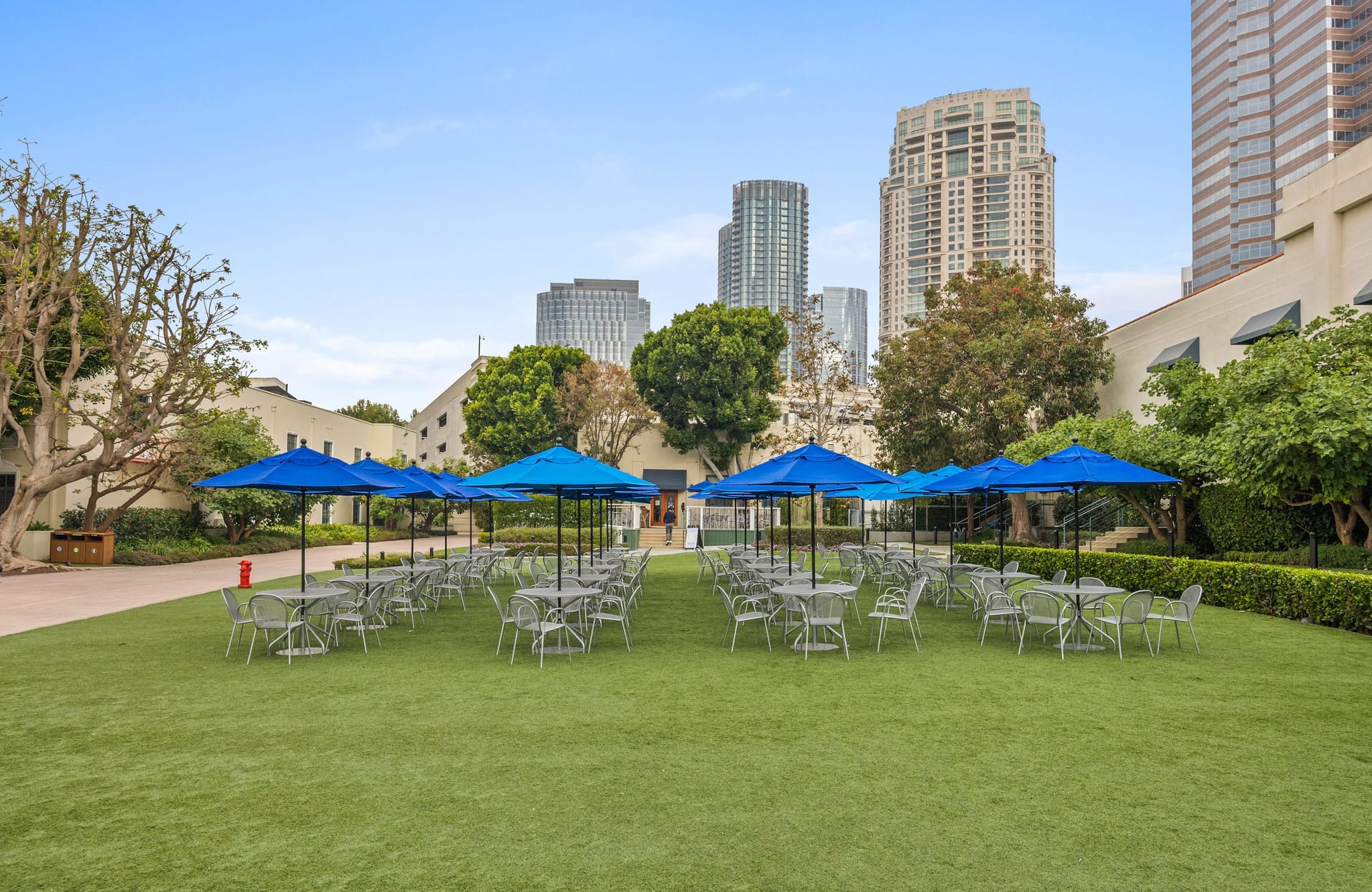 Grassy outdoor seating with blue umbrellas, surrounded by buildings and city skyscrapers.