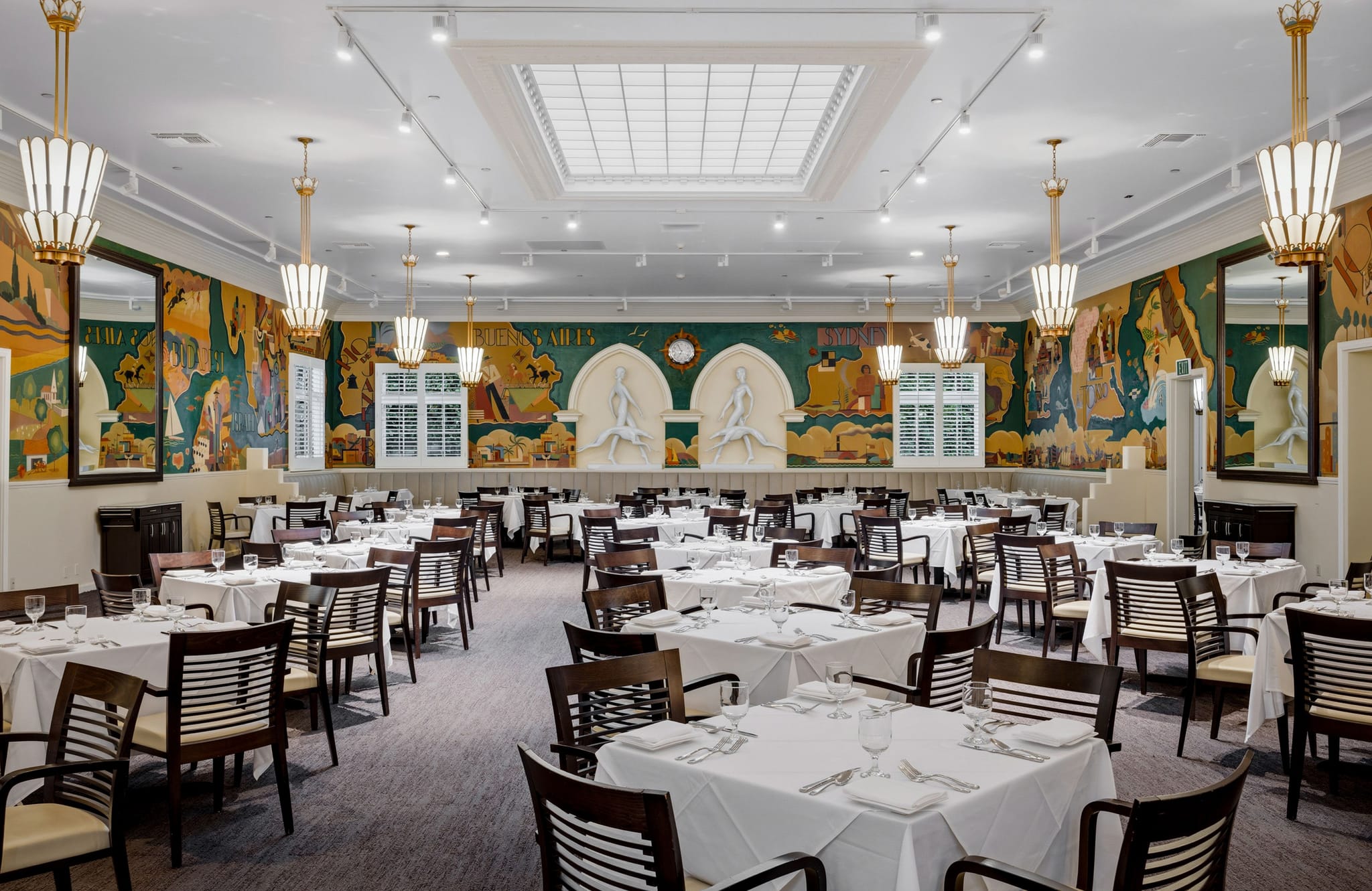 Elegant dining hall with white-clothed tables, colorful wall murals, art deco lights, and a central skylight.