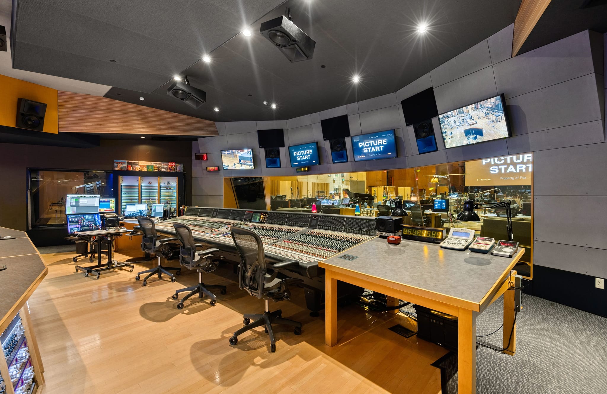 A large, modern post-production audio mixing studio with a long control console, multiple screens displaying "PICTURE START," and sound-dampening walls.