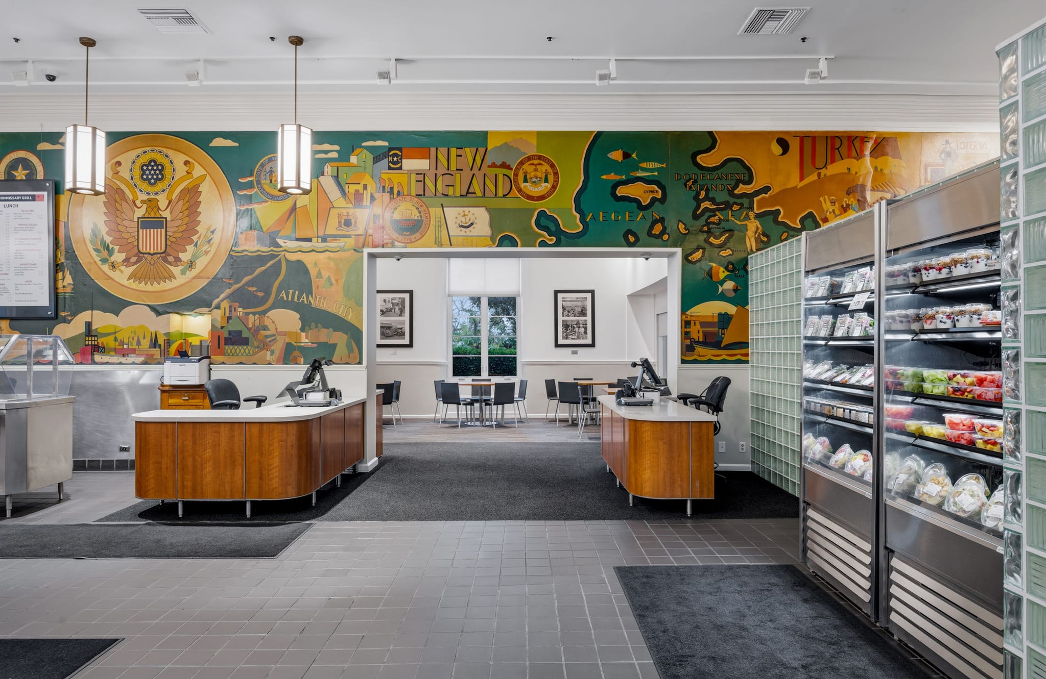 A vibrant dining hall with a large colorful mural depicting historical and geographic themes, featuring service counters and refrigerated grab-and-go food displays.