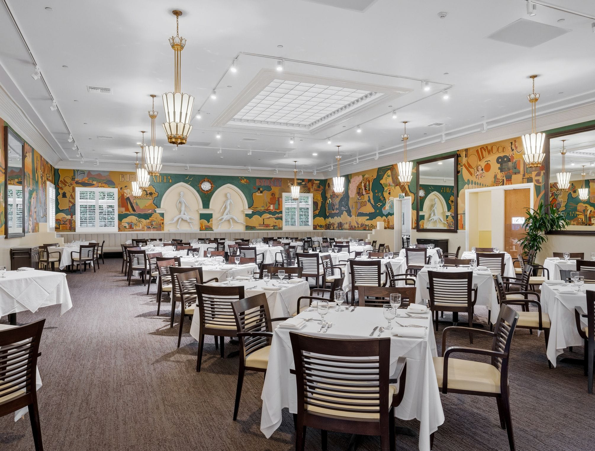 An elegant dining room with white-clothed tables, colorful wall murals, and recessed sculptures.