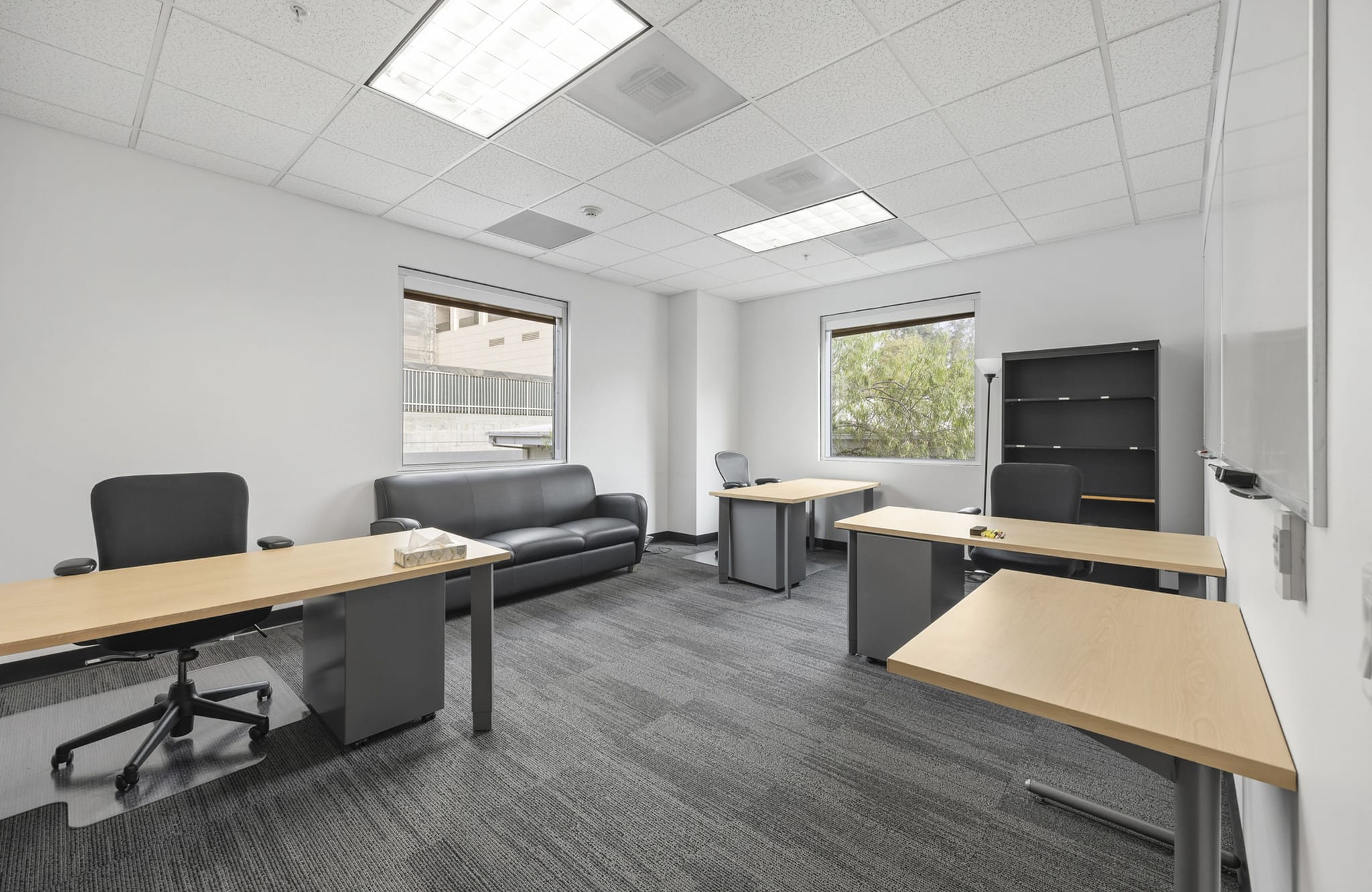A clean office space with several light wood desks, dark office chairs, a black sofa, and a tall black bookshelf on gray carpet.