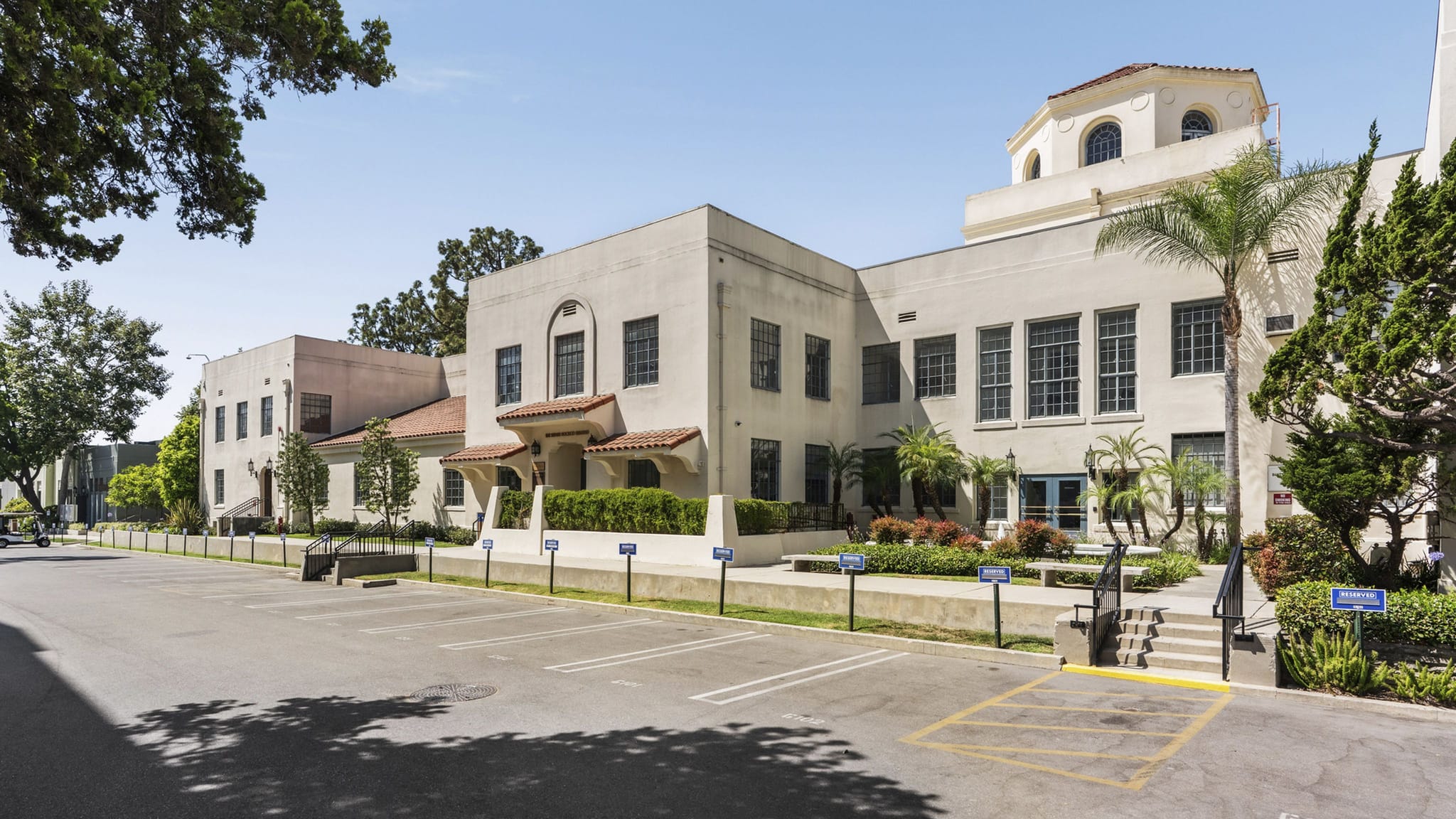 A light-colored Mediterranean Revival building with a prominent cupola, fronted by a parking lot and lush landscaping.