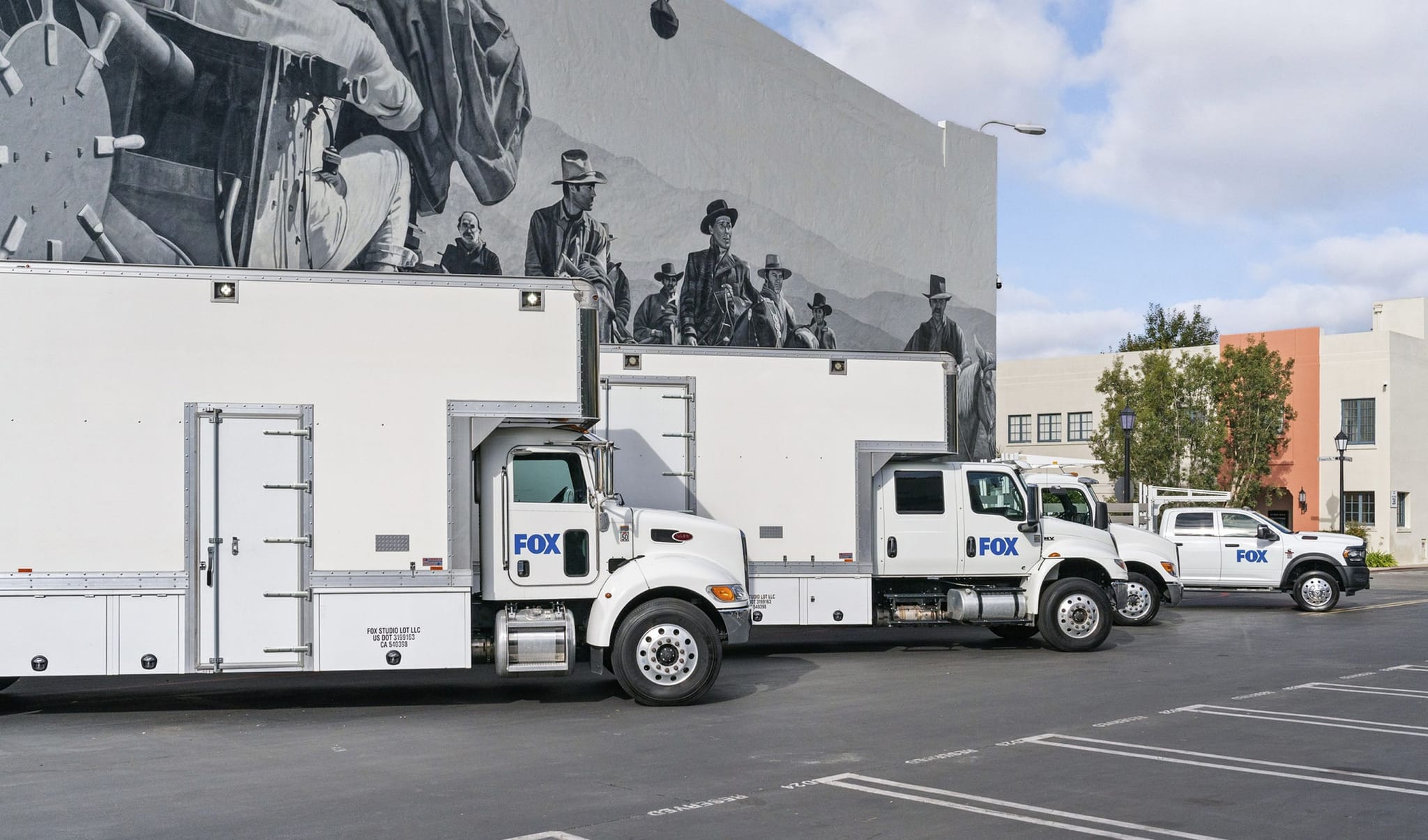 Several white FOX production vehicles parked in front of a building with a black and white mural.