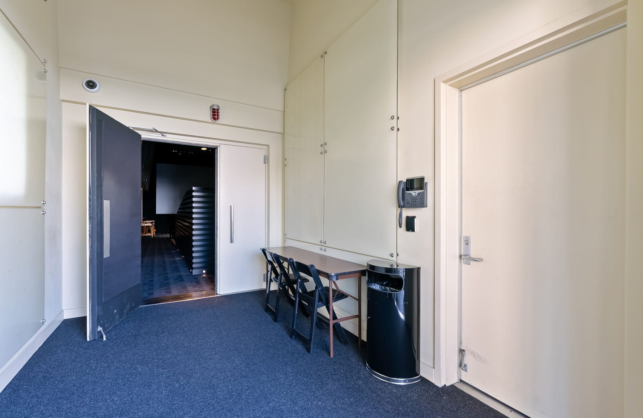 A blue carpeted room with light walls. An open dark door on the left reveals a dark interior with tiered seating. To the right, a table with two black chairs, a wall phone, and a trash can stands beside a closed door.