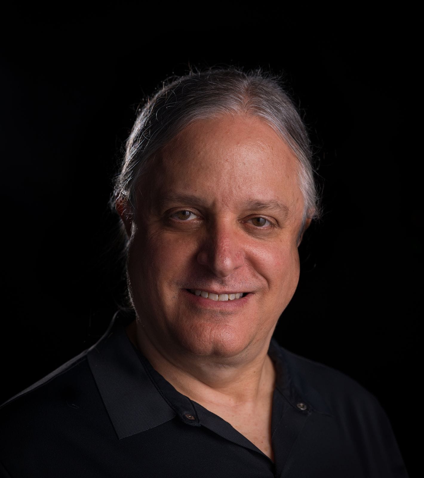 Smiling man with gray hair in a black shirt against a dark background.