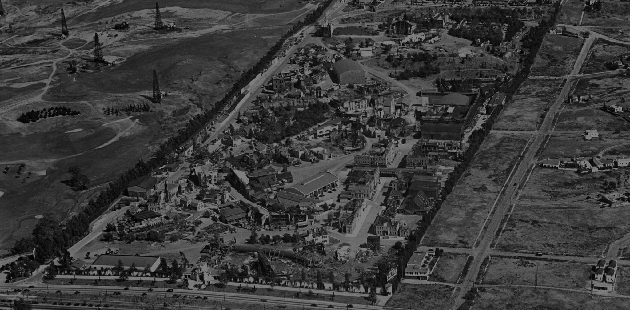 Aerial black and white view of a large studio lot or entertainment complex next to hills with oil derricks.