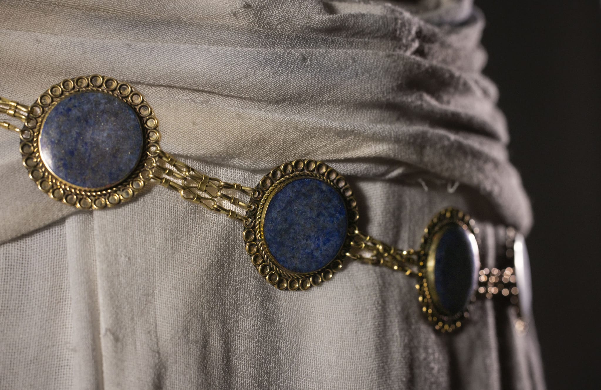 A gold-toned belt with circular lapis lazuli stones worn over light, draped fabric.